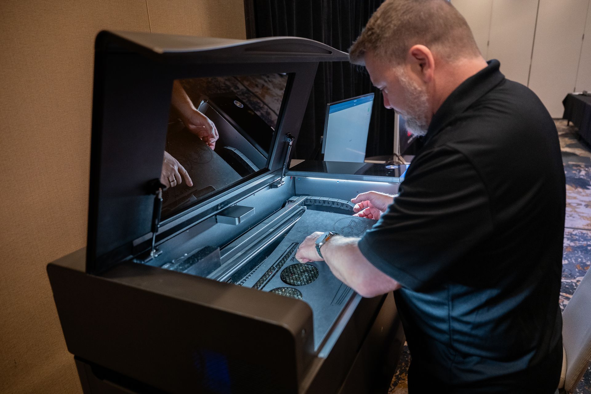 Man working on a dark-colored laser cutter machine indoors. He is wearing a black shirt and focused.