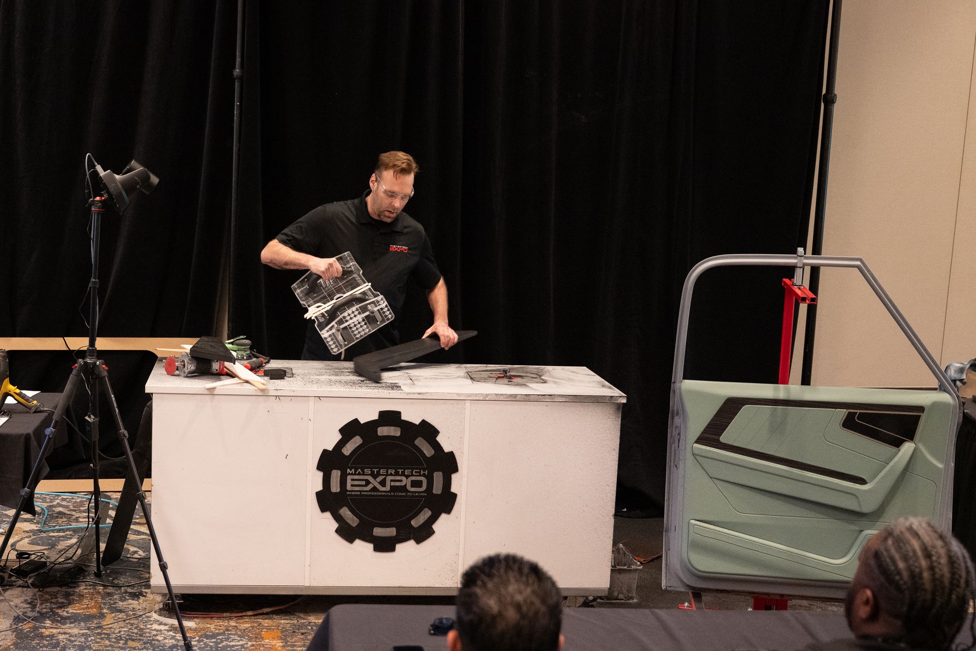 Man demonstrating vehicle door panel work at an event. White table, black backdrop, vehicle door in frame.