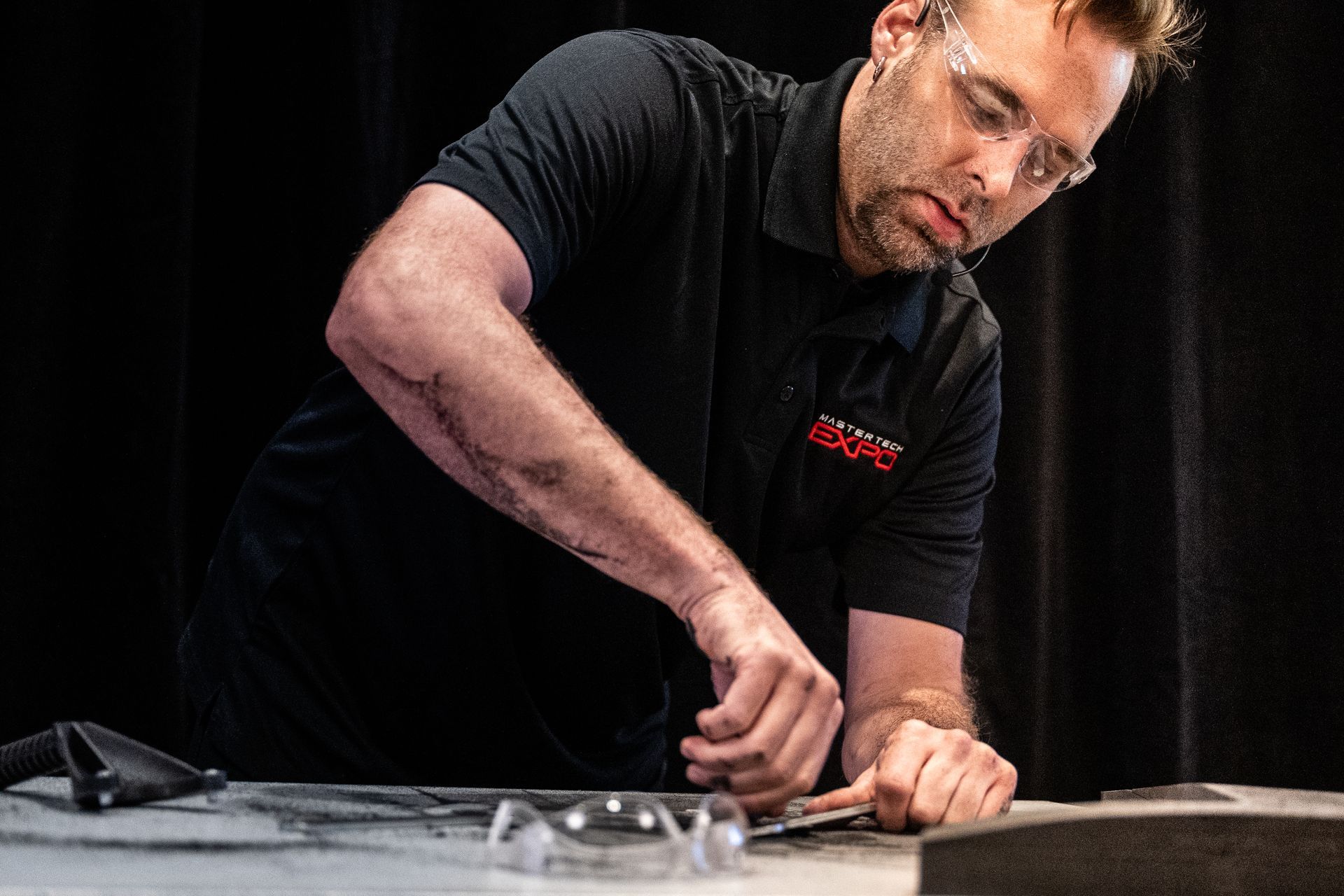 Man in black shirt and safety glasses working on a surface; holding a tool.