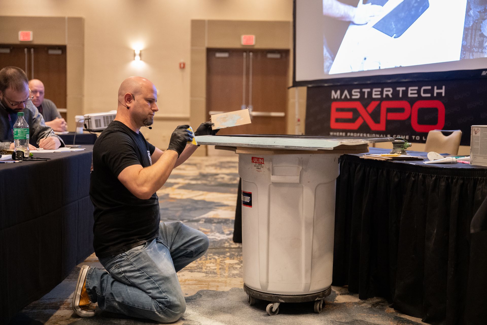 Man in black shirt kneels, working on a surface atop a trash can at a conference, the Mastertech Expo.