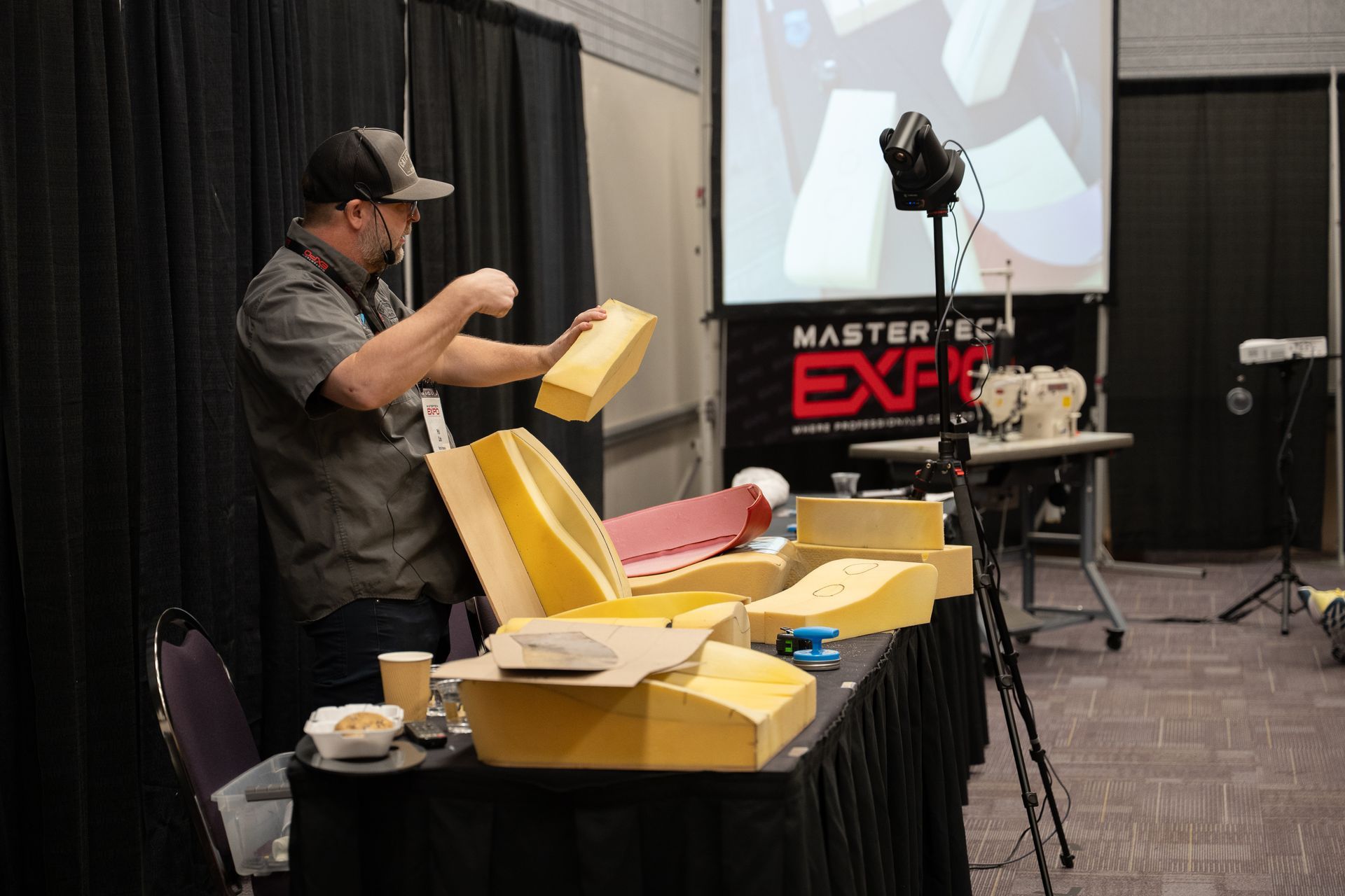 Man demonstrating foam shapes at a convention booth with a projector screen in the background.