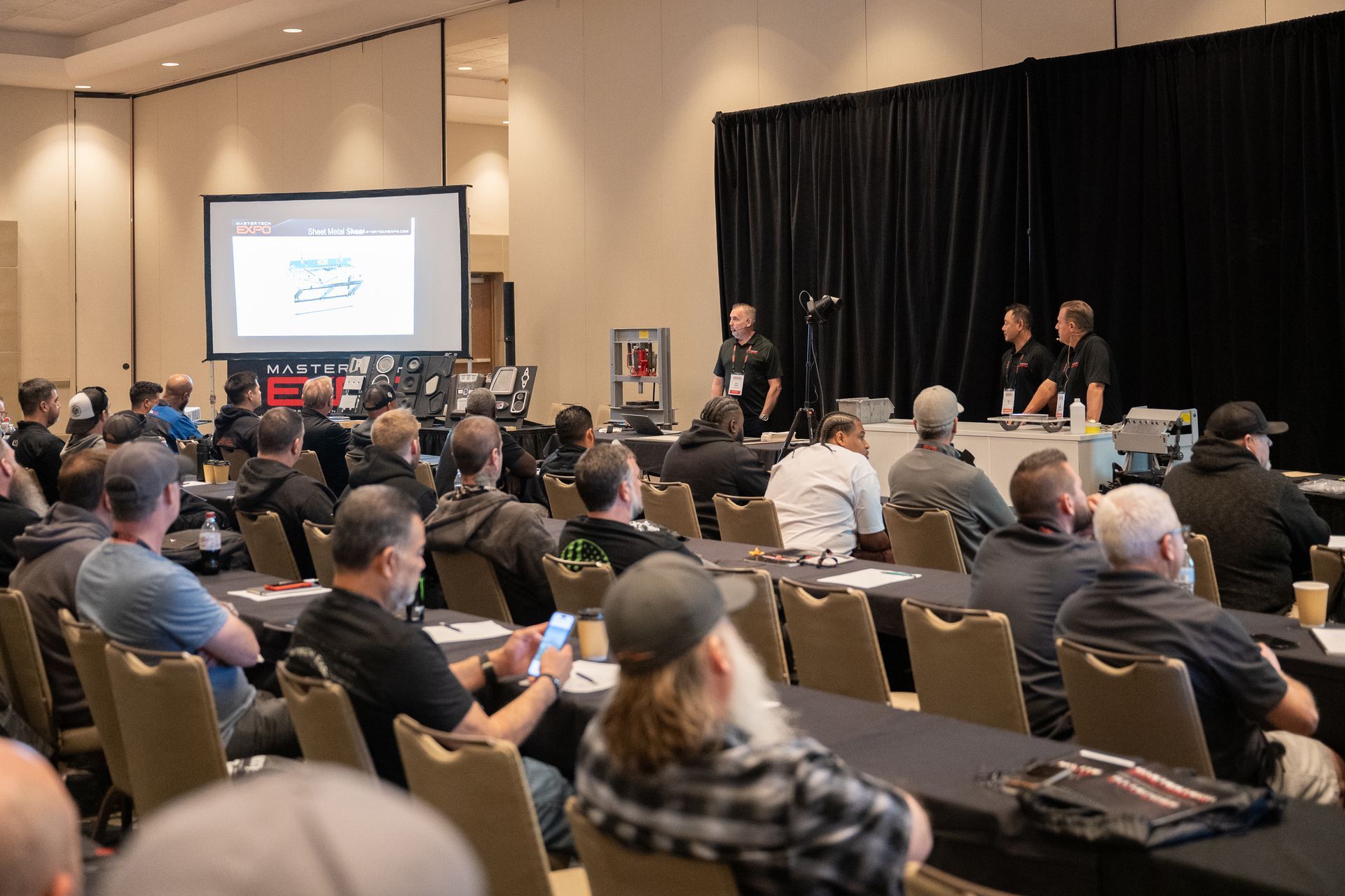 Conference presentation: Speakers on stage, audience seated at tables, projected image.
