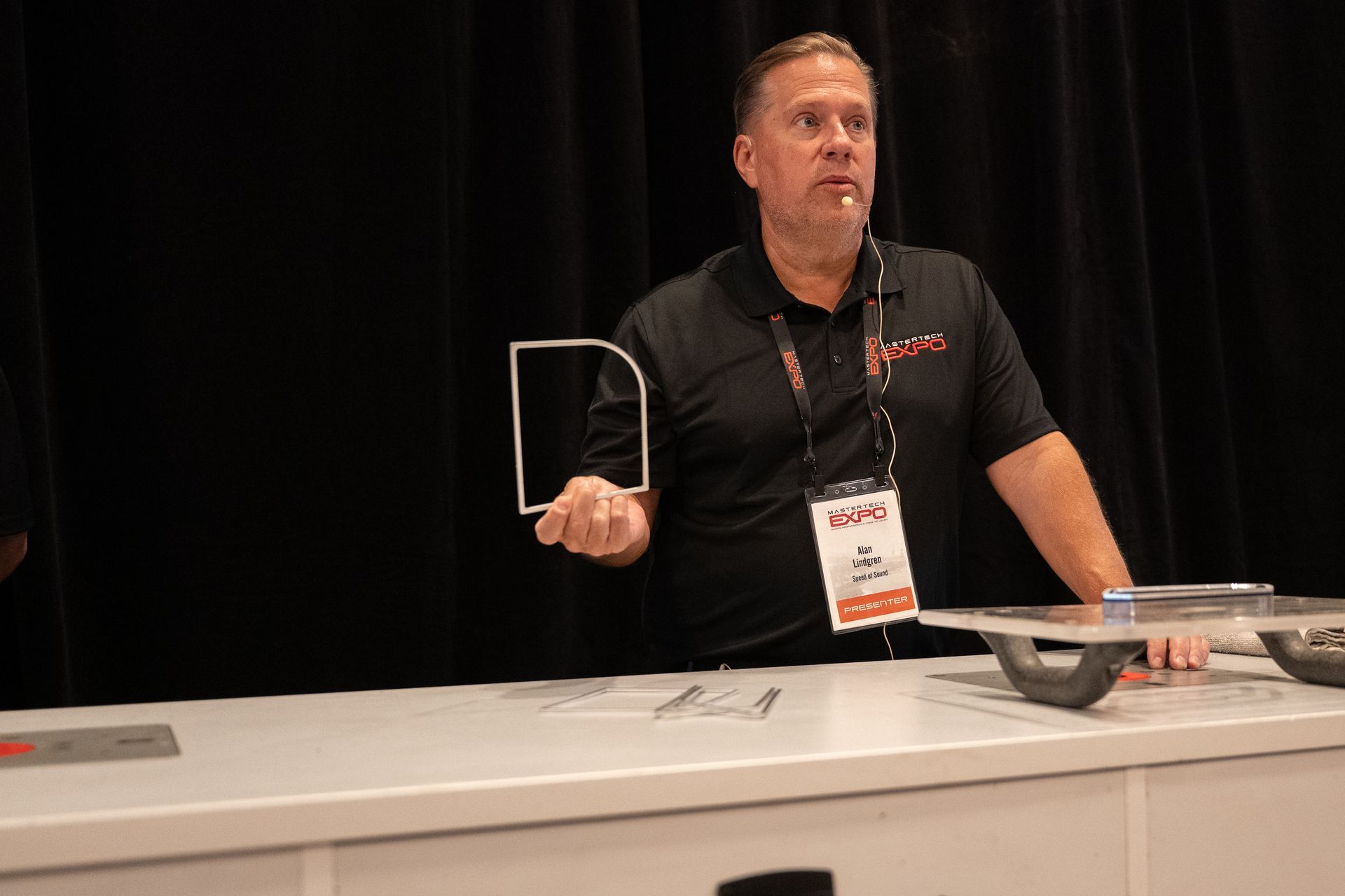 Man at a trade show booth holds a clear, angled plastic frame; black backdrop and white counter.