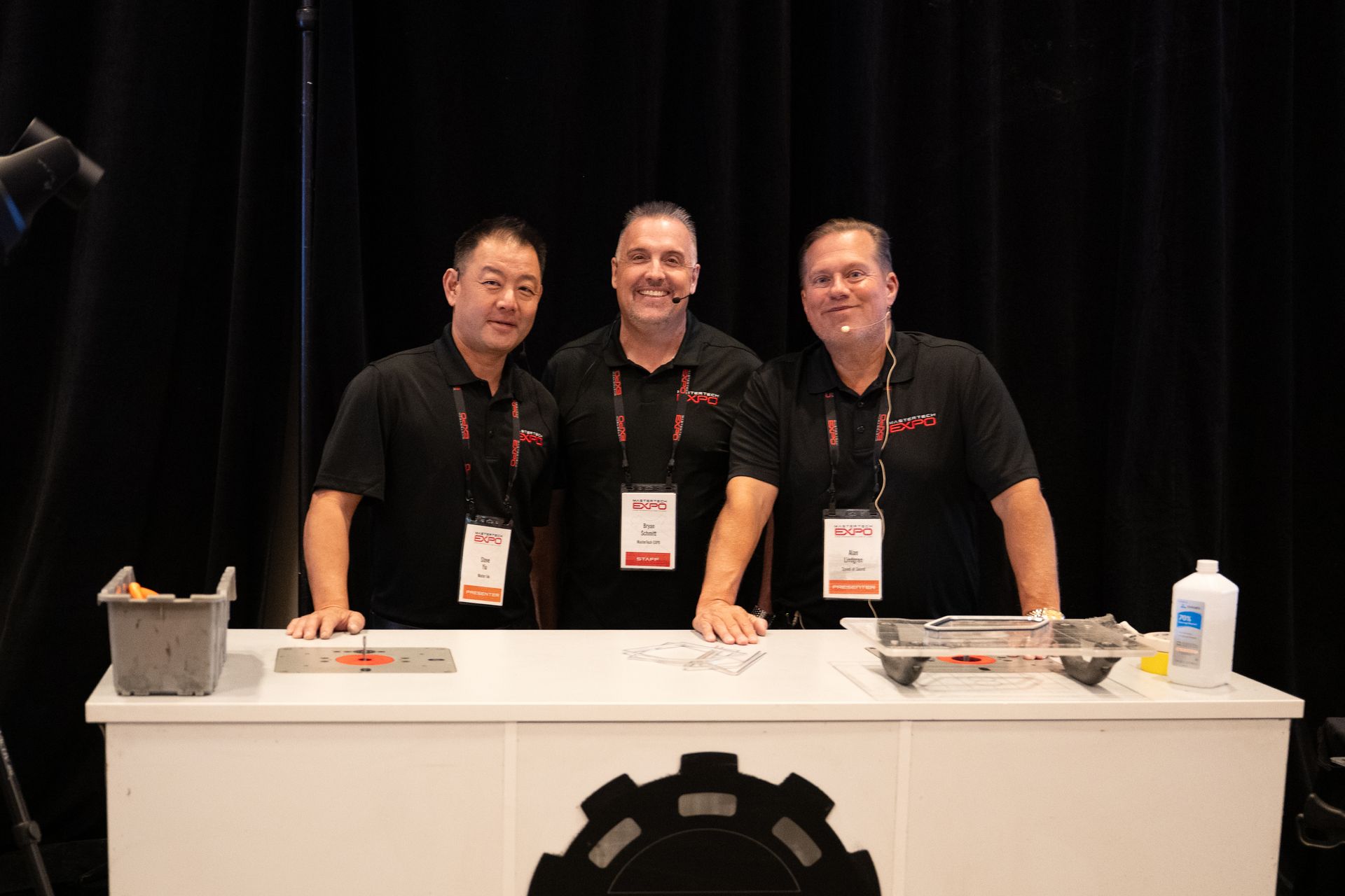 Three men smiling behind a booth at an event. They wear black shirts and lanyards. A table with tools and supplies is in front of them.