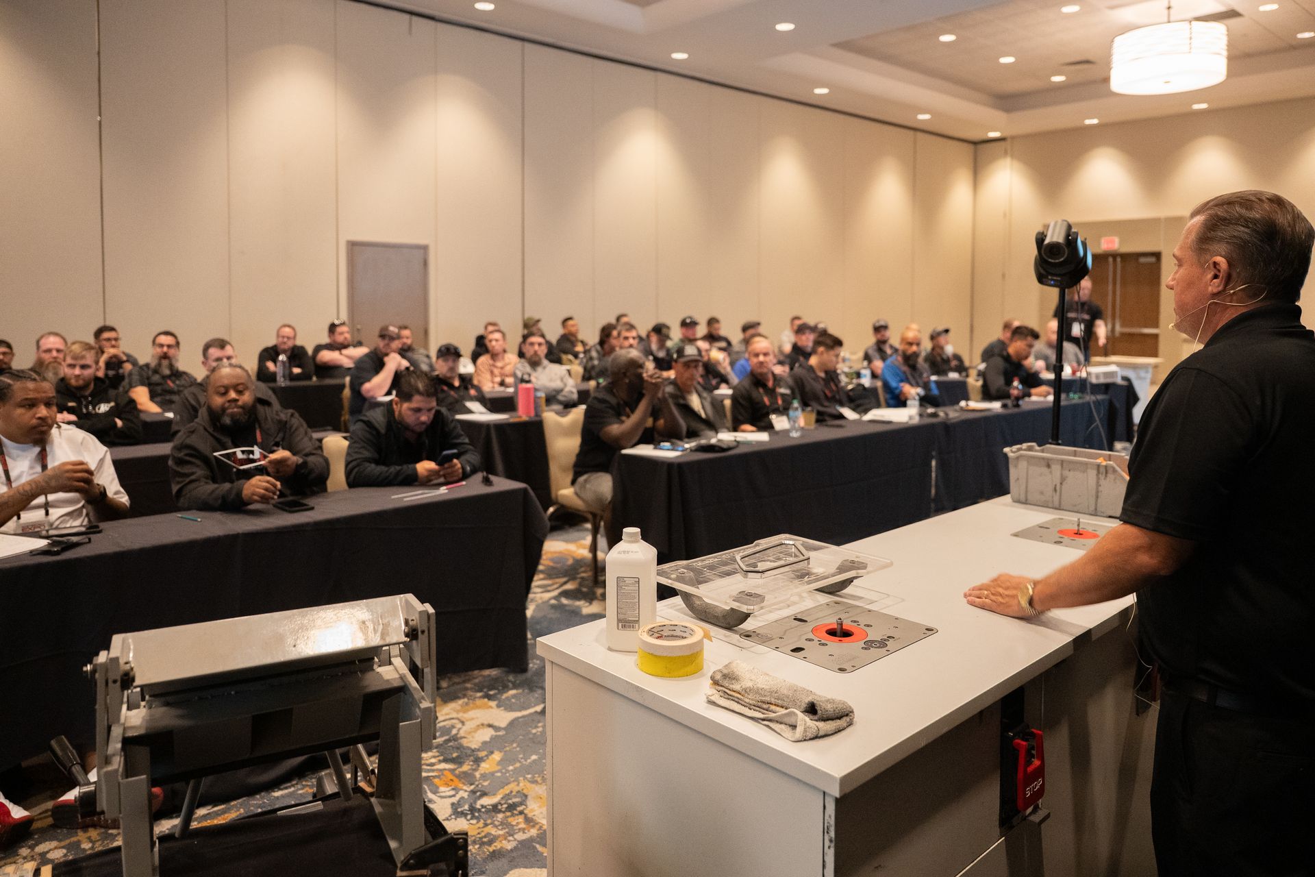 A speaker at a seminar stands before a crowd, demonstrating with items on a table.