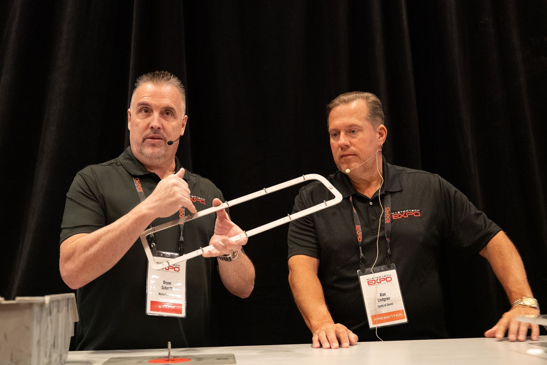 Two men in black shirts demonstrate a tool, one holding it up, at a conference booth.