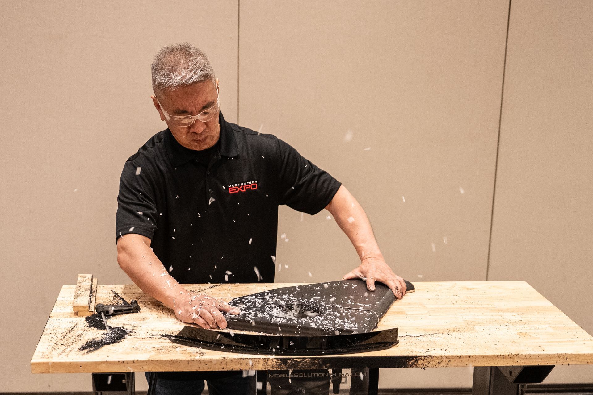 Man using a tool to scrape a black object on a wooden table, debris in air.
