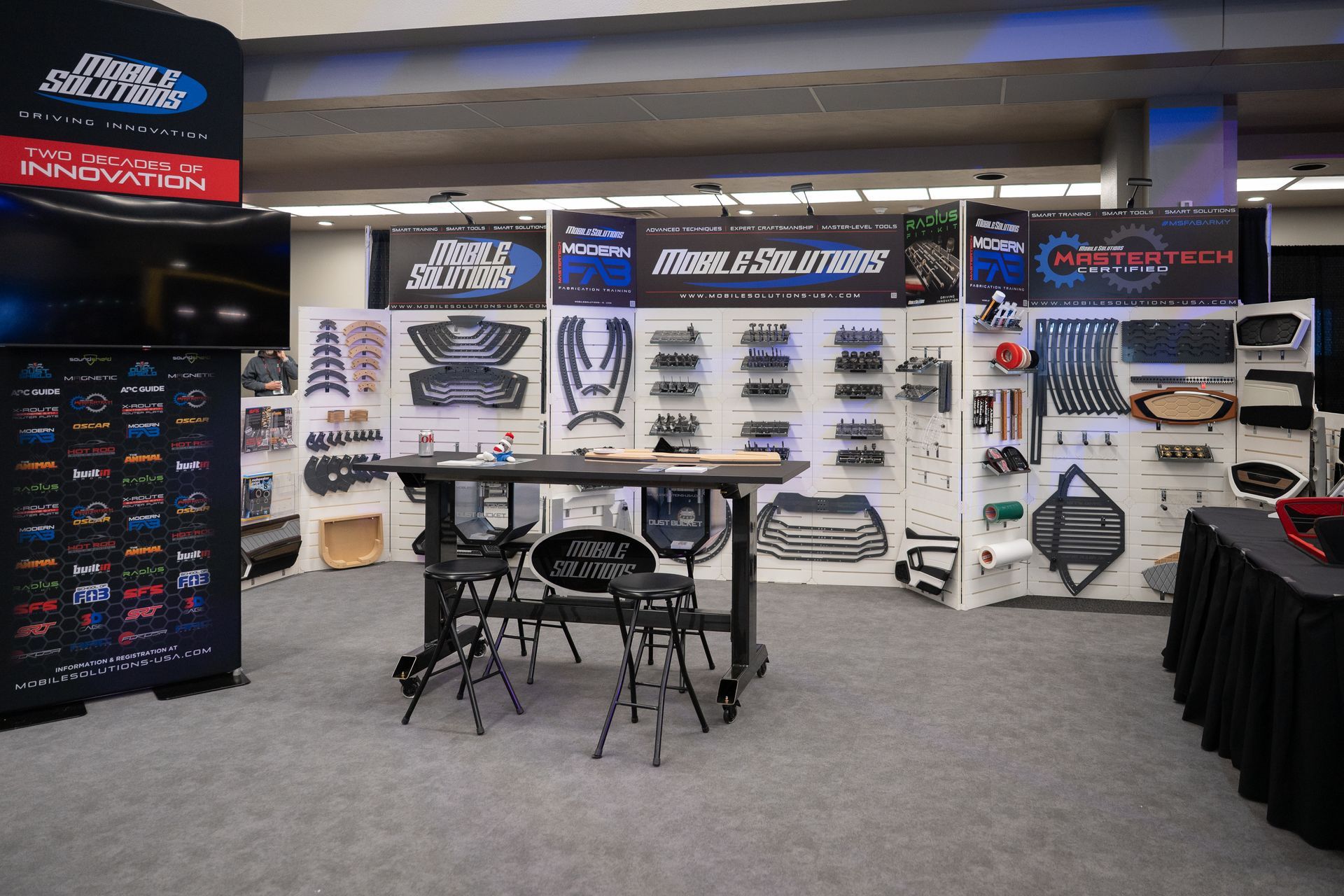 Trade show booth displaying automotive parts. Black, gray, and blue color scheme. Parts are mounted on white panels. Table and stools in center.