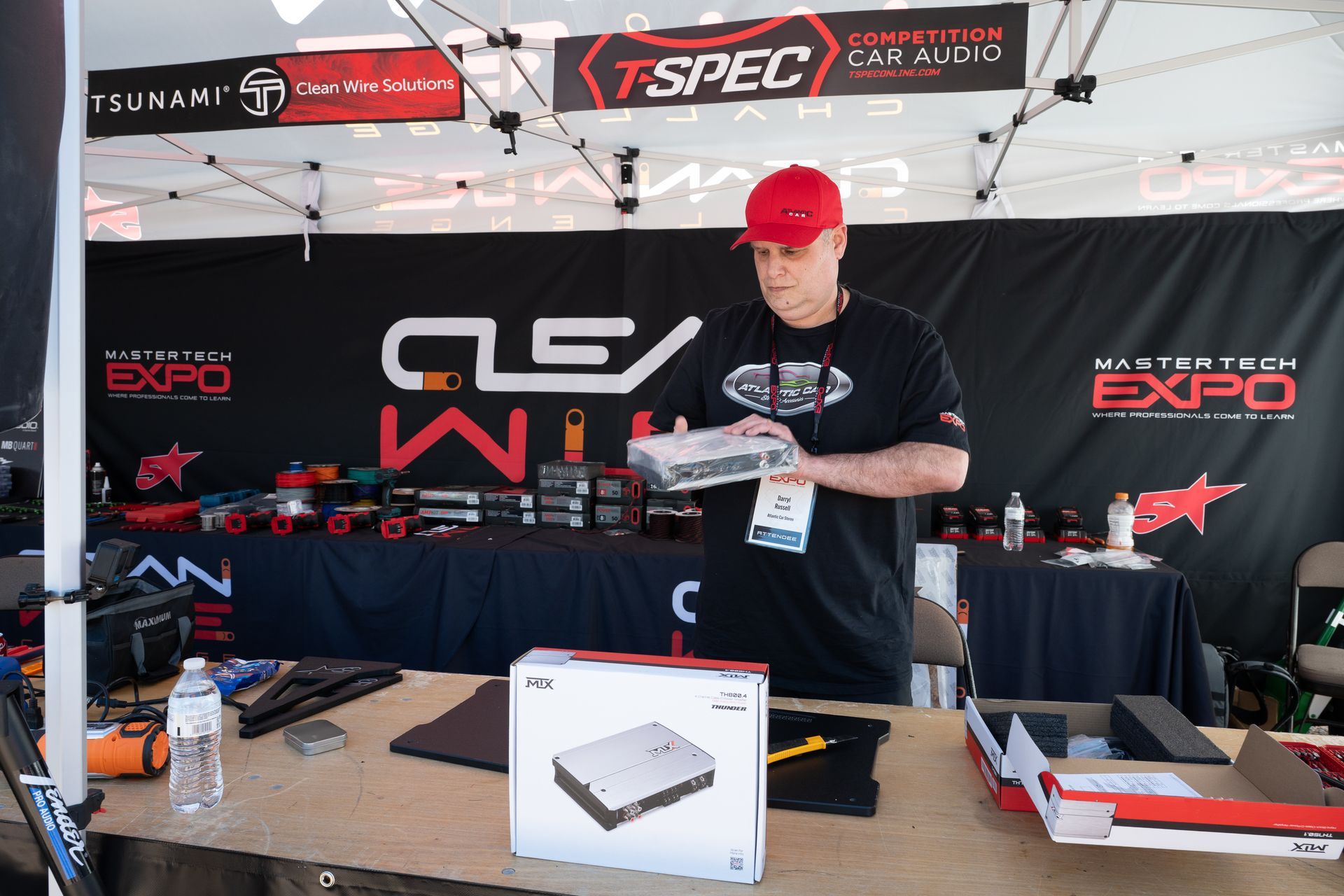 Man at a vendor booth holding a product box, surrounded by audio equipment and expo banner.