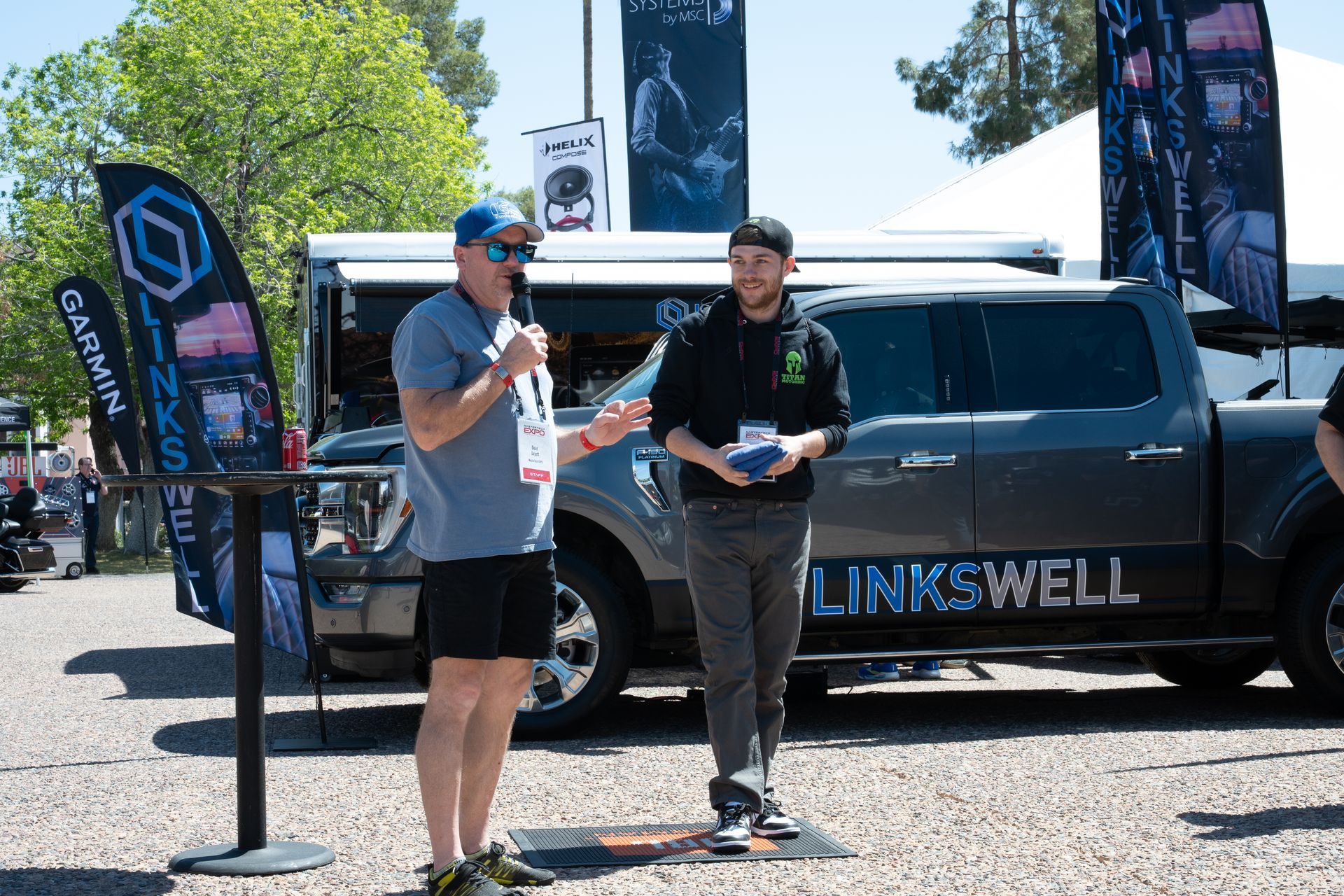 Man speaking into a microphone, presenting an award to another man near a truck at an outdoor event.