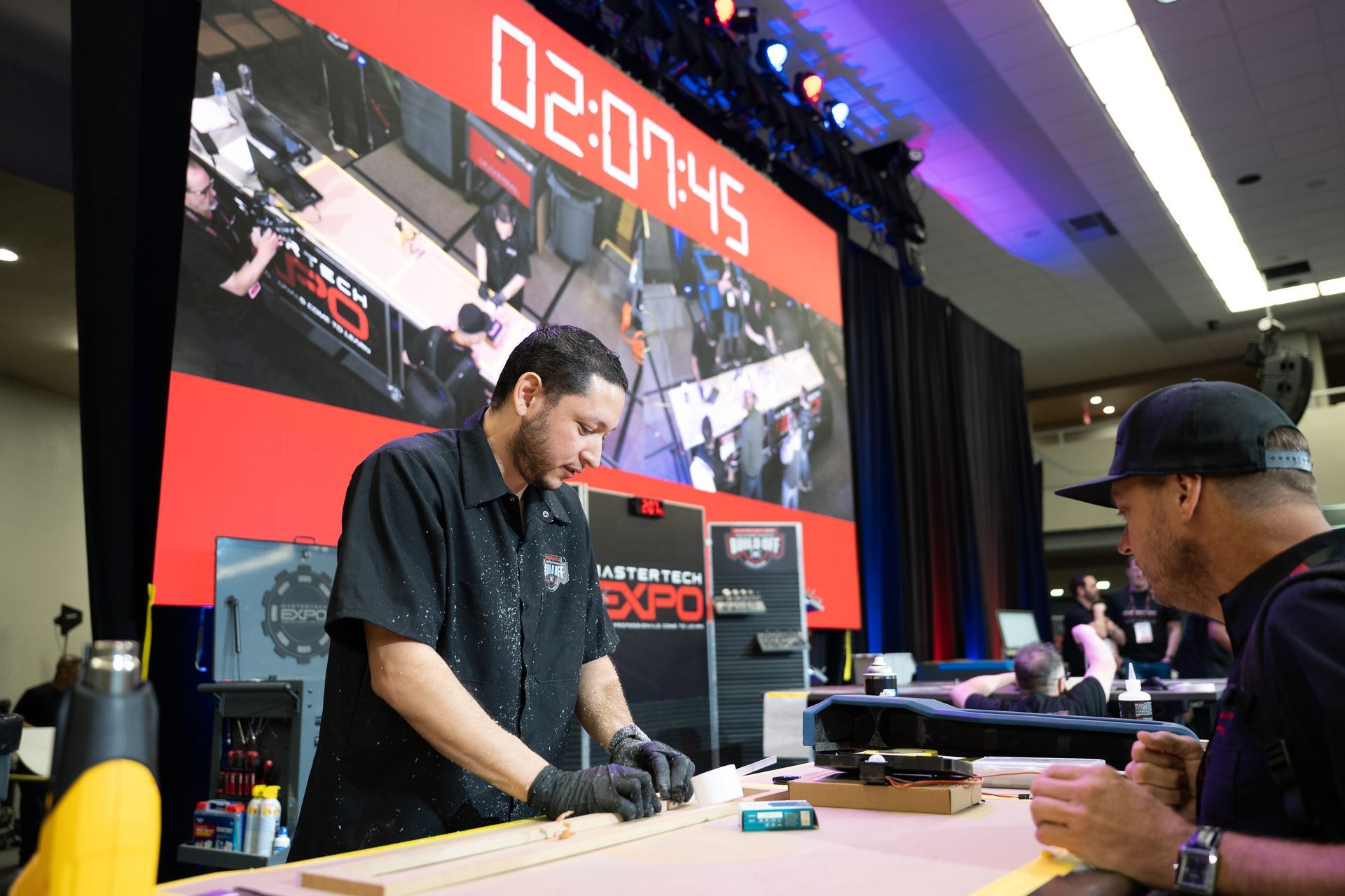 Man works at a table, with another man observing. Overhead timer shows 02:07:45 at an expo.