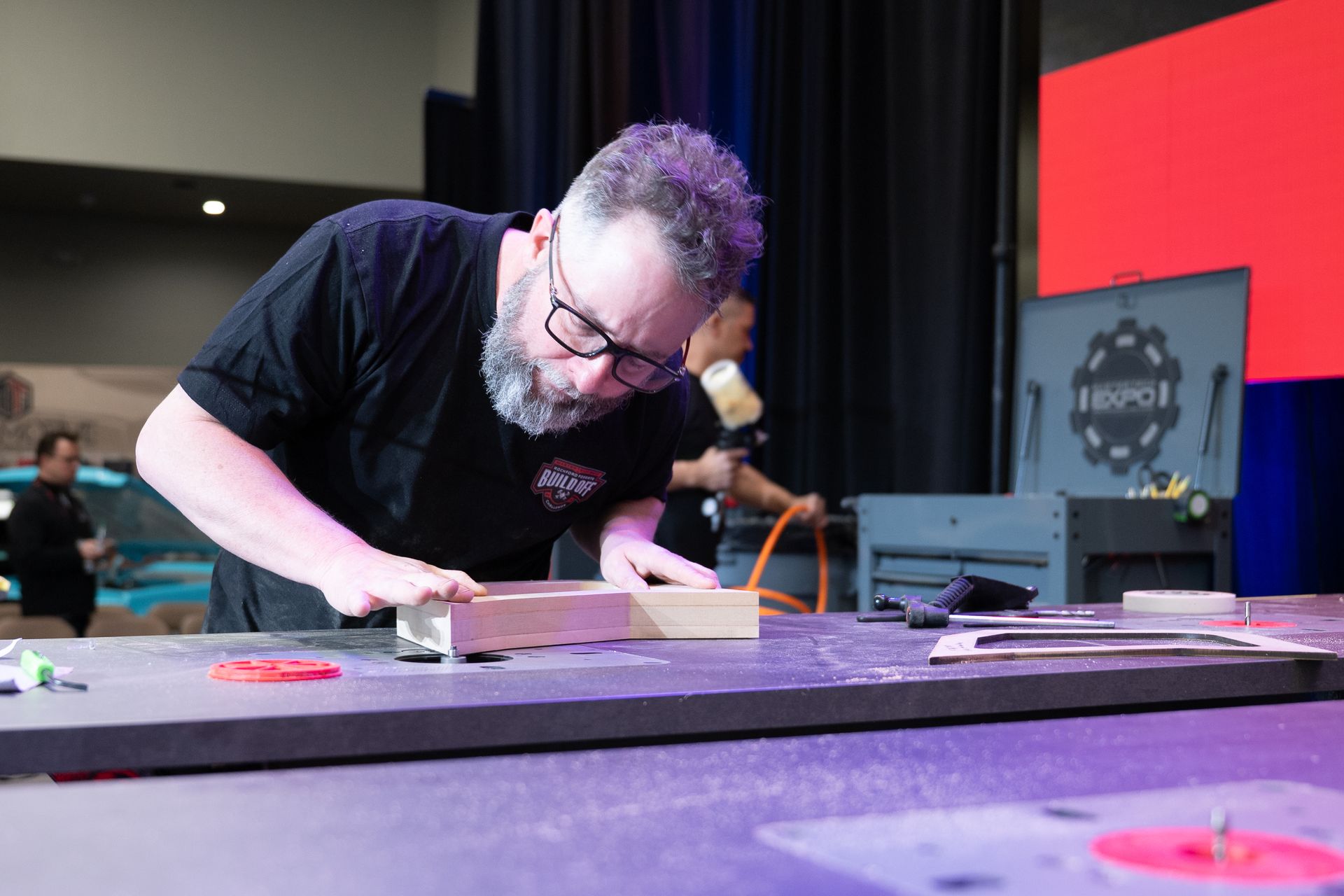 Man with glasses working on wood at a workbench. Black shirt, focused expression, indoors.