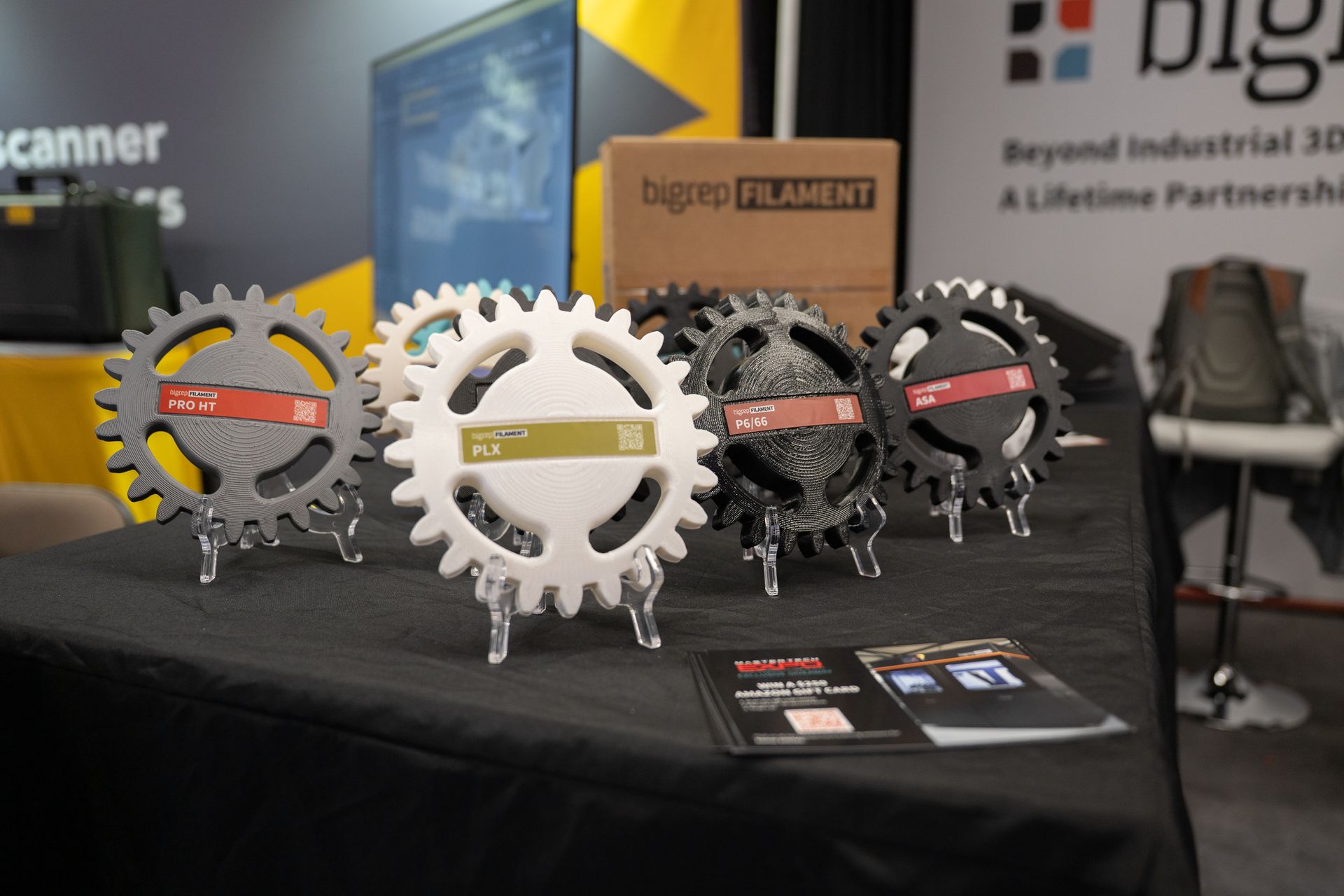 Display of 3D-printed gears with product labels on a table at a trade show.