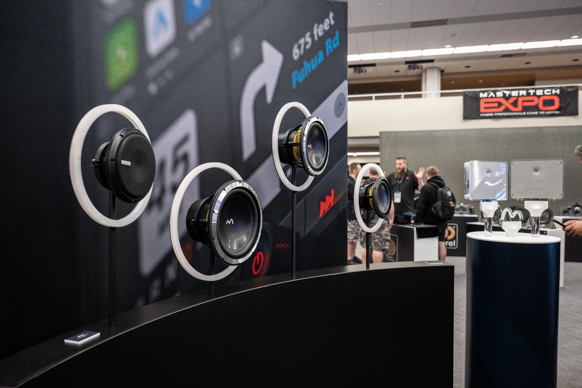 Speakers displayed at an expo. People in the background. Black and silver with white rings.