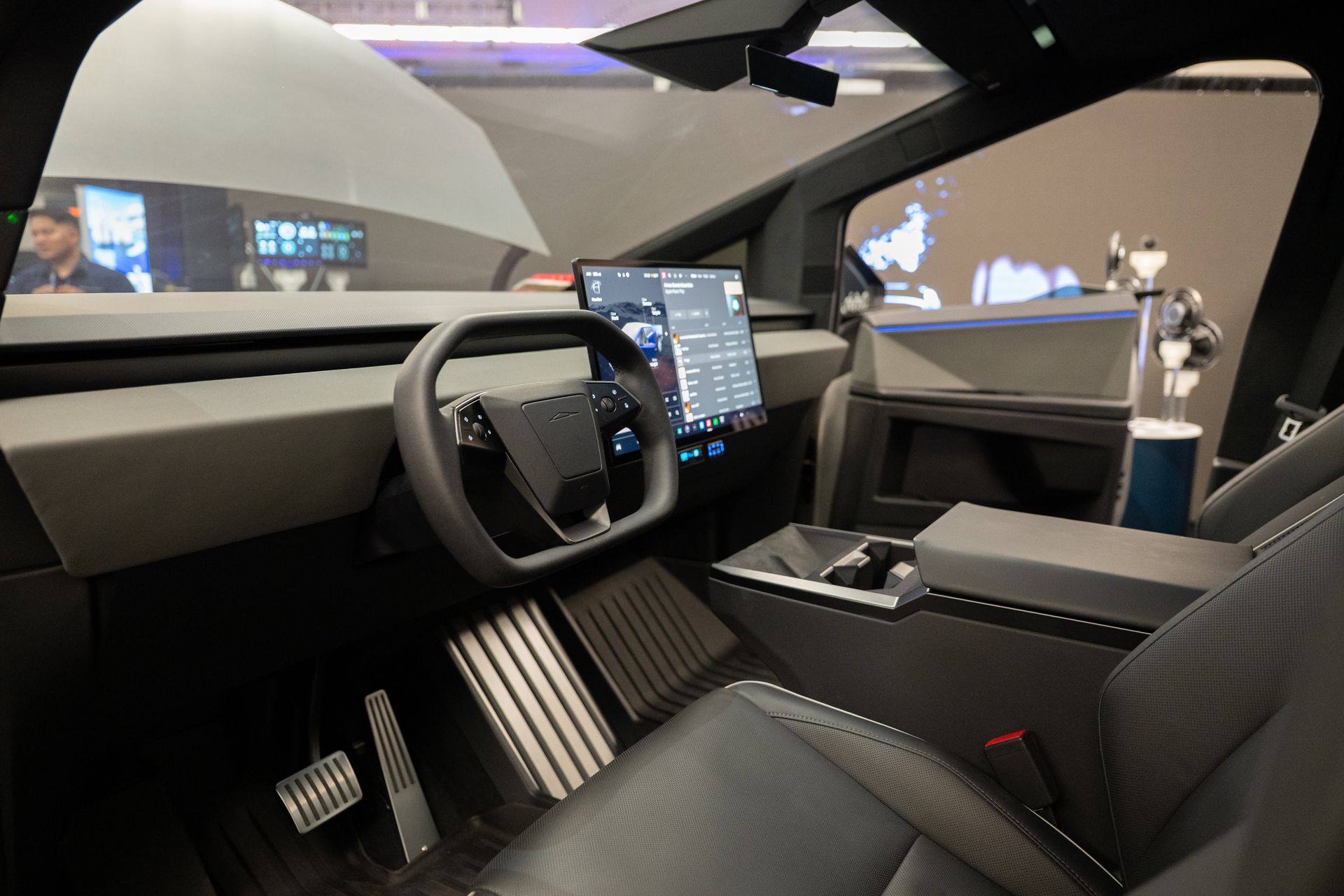Interior view of a Tesla Cybertruck with gray dashboard, black steering wheel, and large central touchscreen.