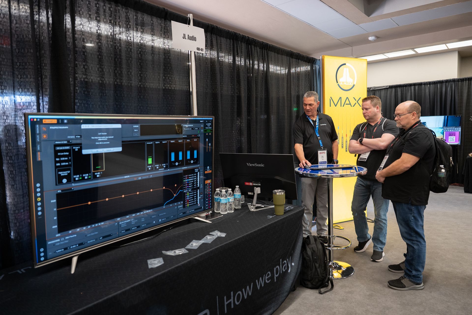 Three men at a trade show booth, looking at a large screen displaying software interface.