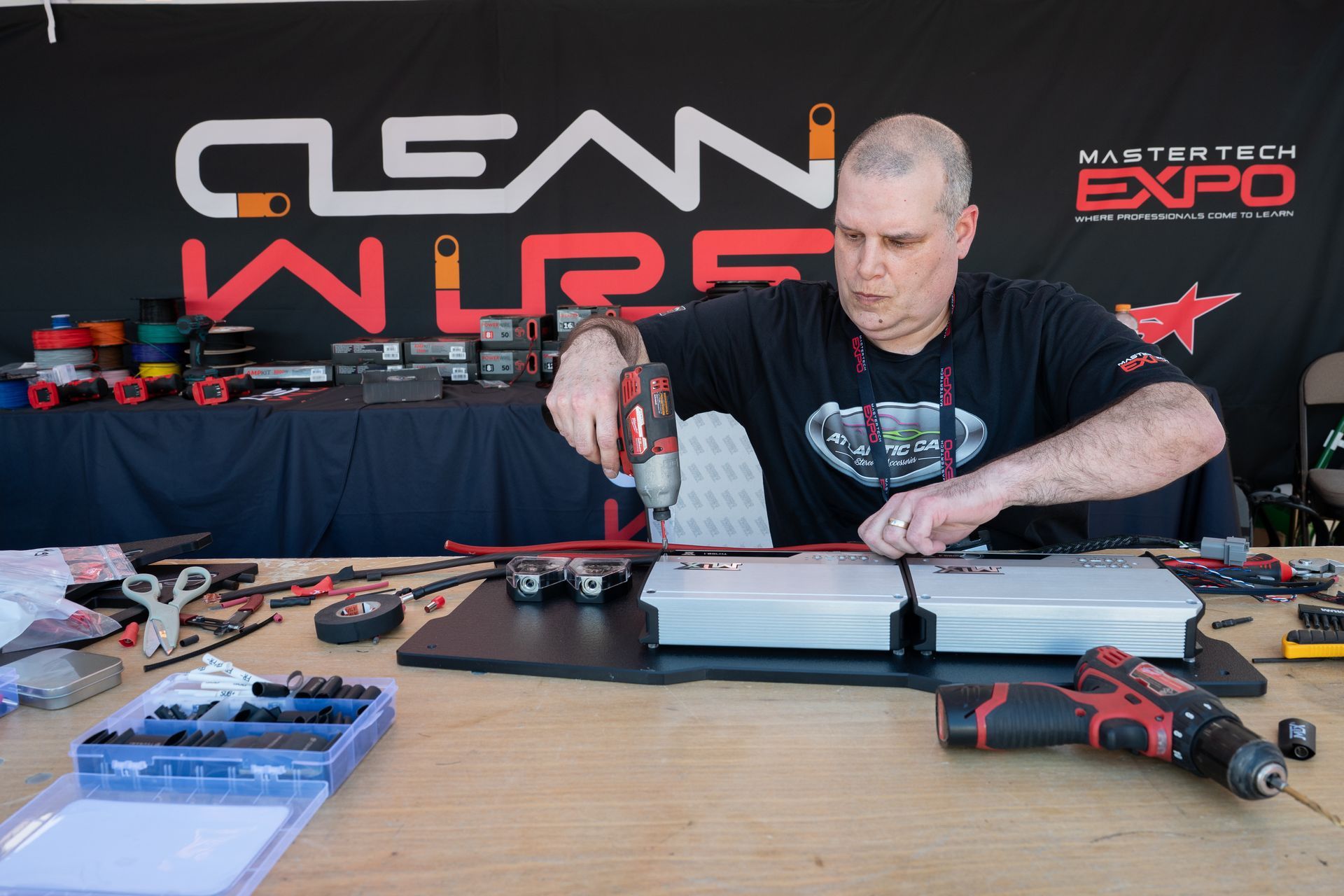 Man using a drill on a car audio setup at a trade show booth with Clean Wire and Mastertech Expo branding.