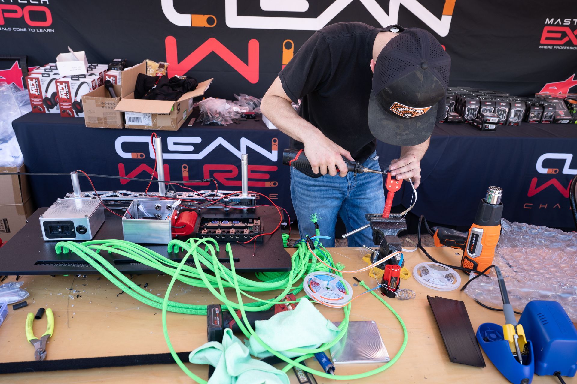 Man working on electrical components at a table, soldering and using tools. Green wires, black table, logo 