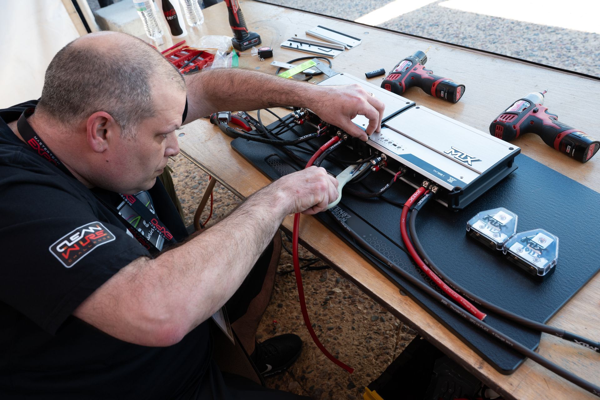Man wiring a car audio amplifier on a wooden table, two power drills nearby.