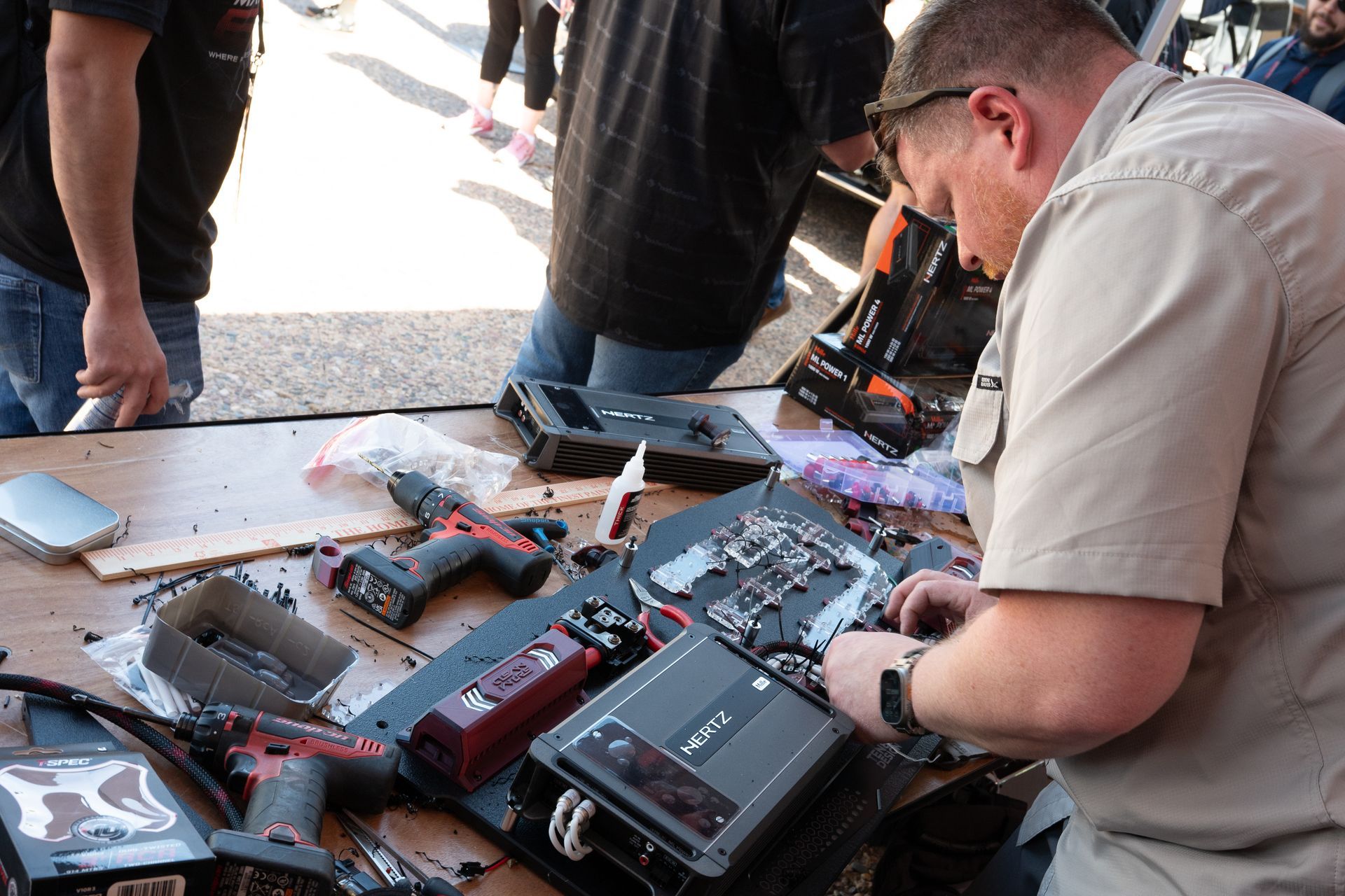 Person in khaki shirt dismantles electronics at outdoor table, surrounded by tools and components.