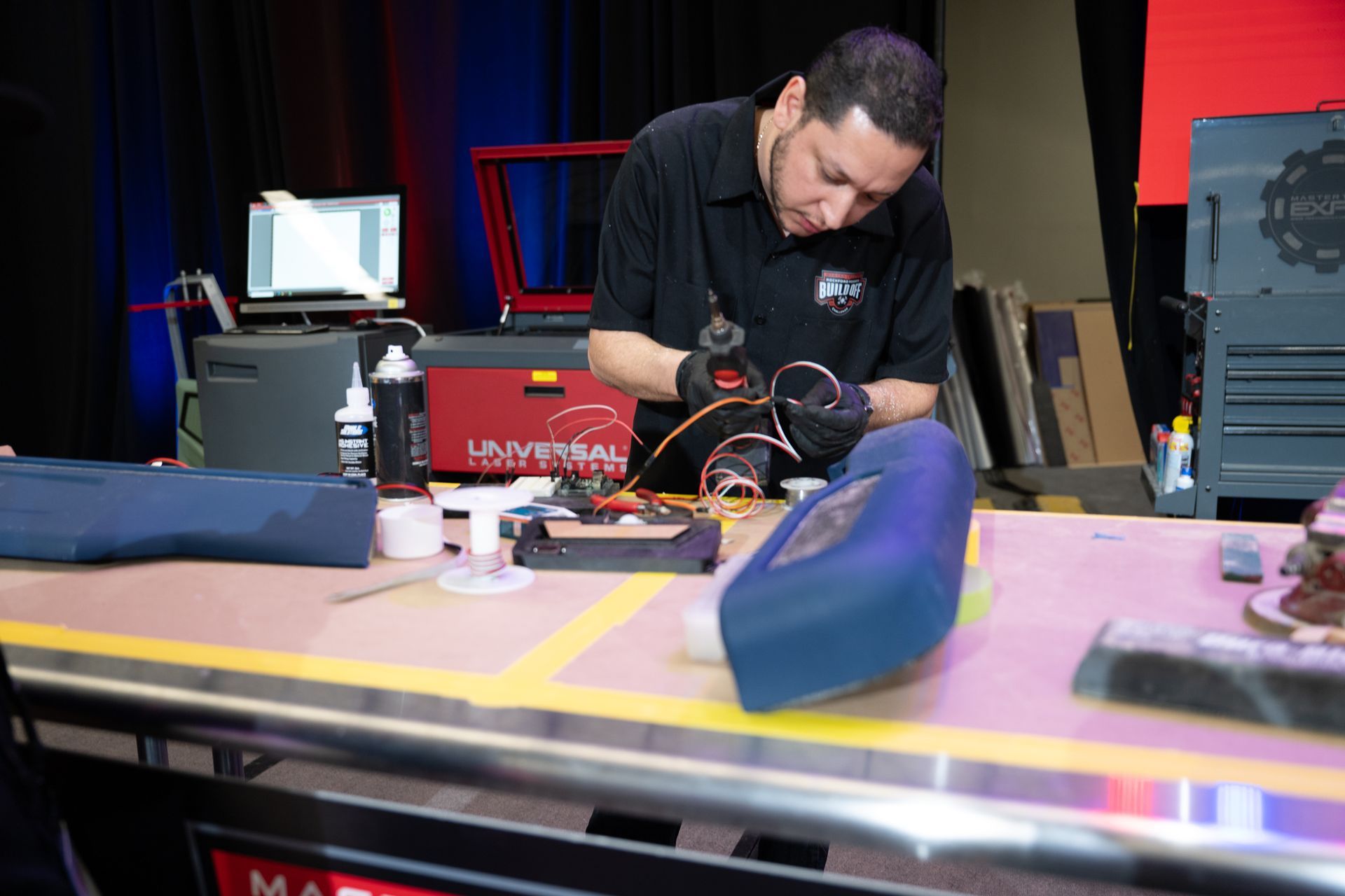 Man working on electronics at a workbench with equipment, tools, and a blue car part.