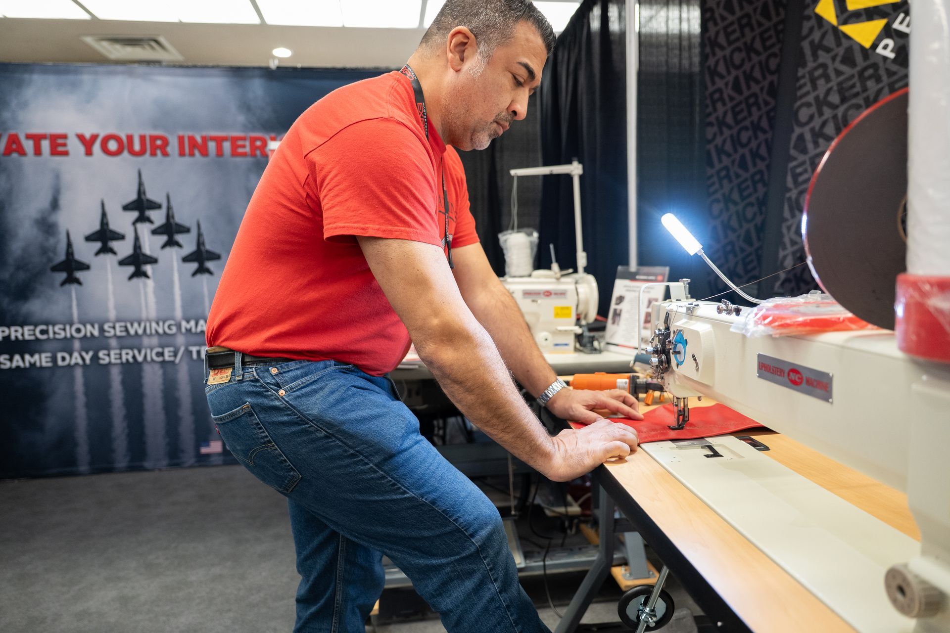 Man in red shirt sews leather at a workshop. He is bent over the sewing machine.