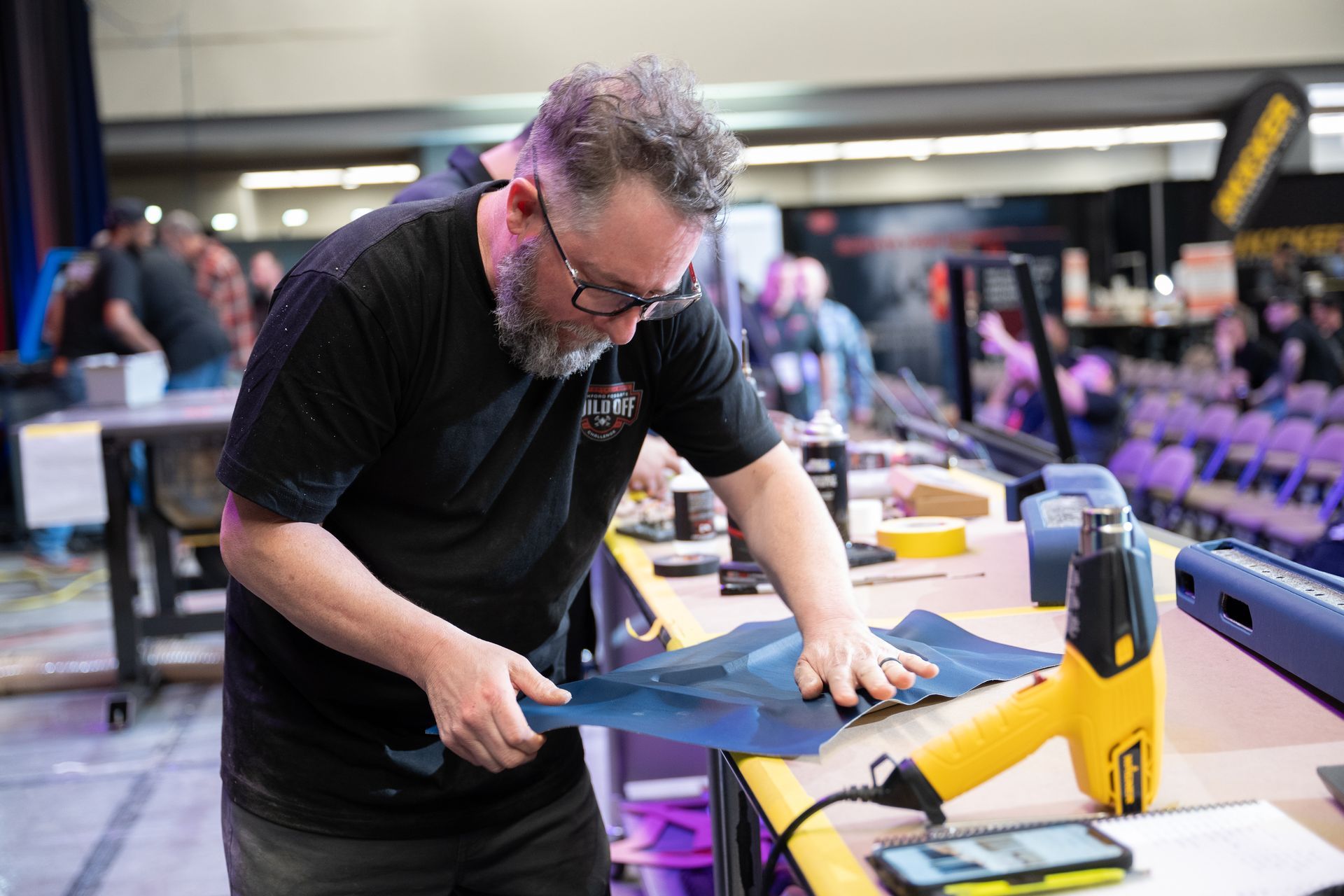 Man working with a blue material at a table, using a heat gun. Indoors, convention or trade show setting.