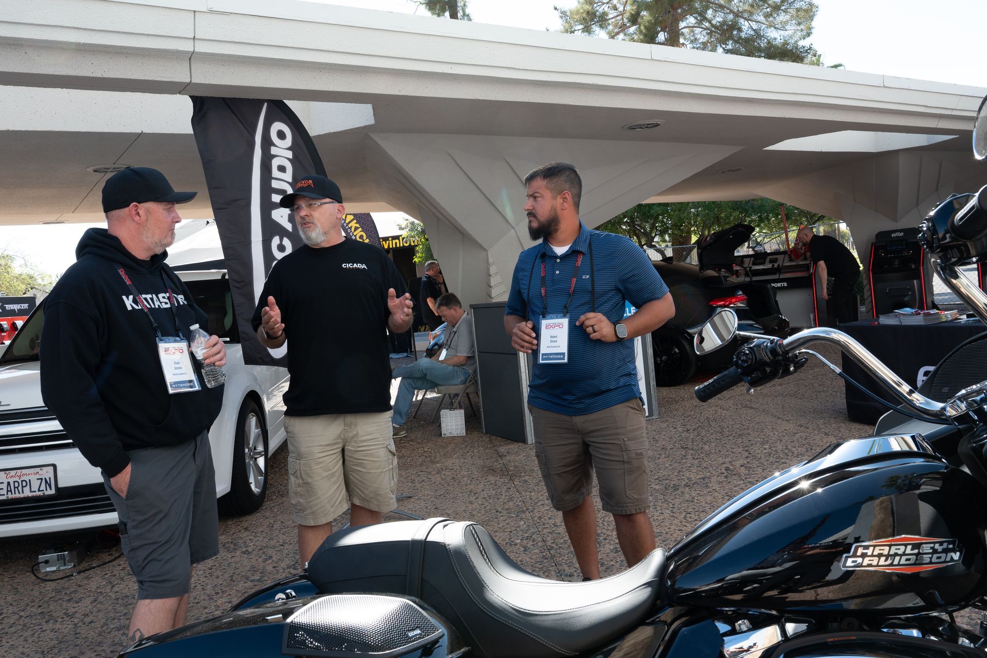 Three men talking near a Harley-Davidson motorcycle at an outdoor event, under a white structure.