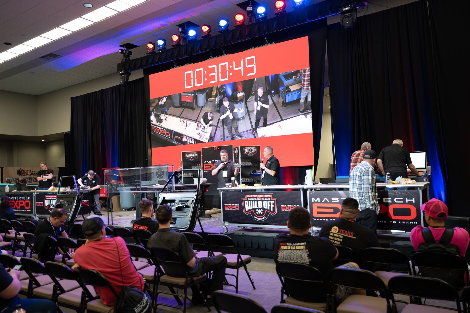 A tech competition with participants at work stations, a large screen displaying the timer and an audience seated in chairs.