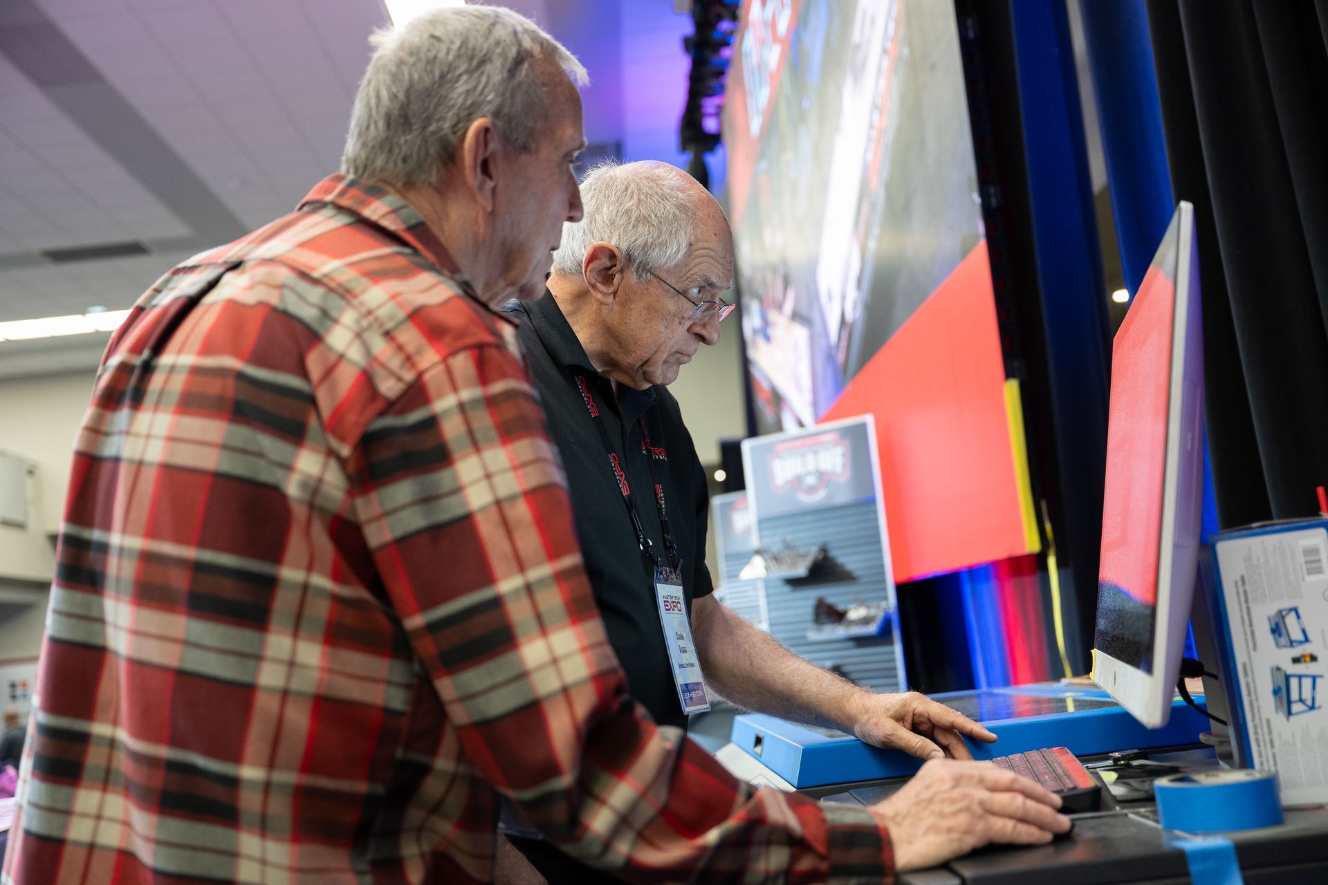 Two older men looking at a computer screen, one using controls at a booth. Red plaid shirt and black shirt.