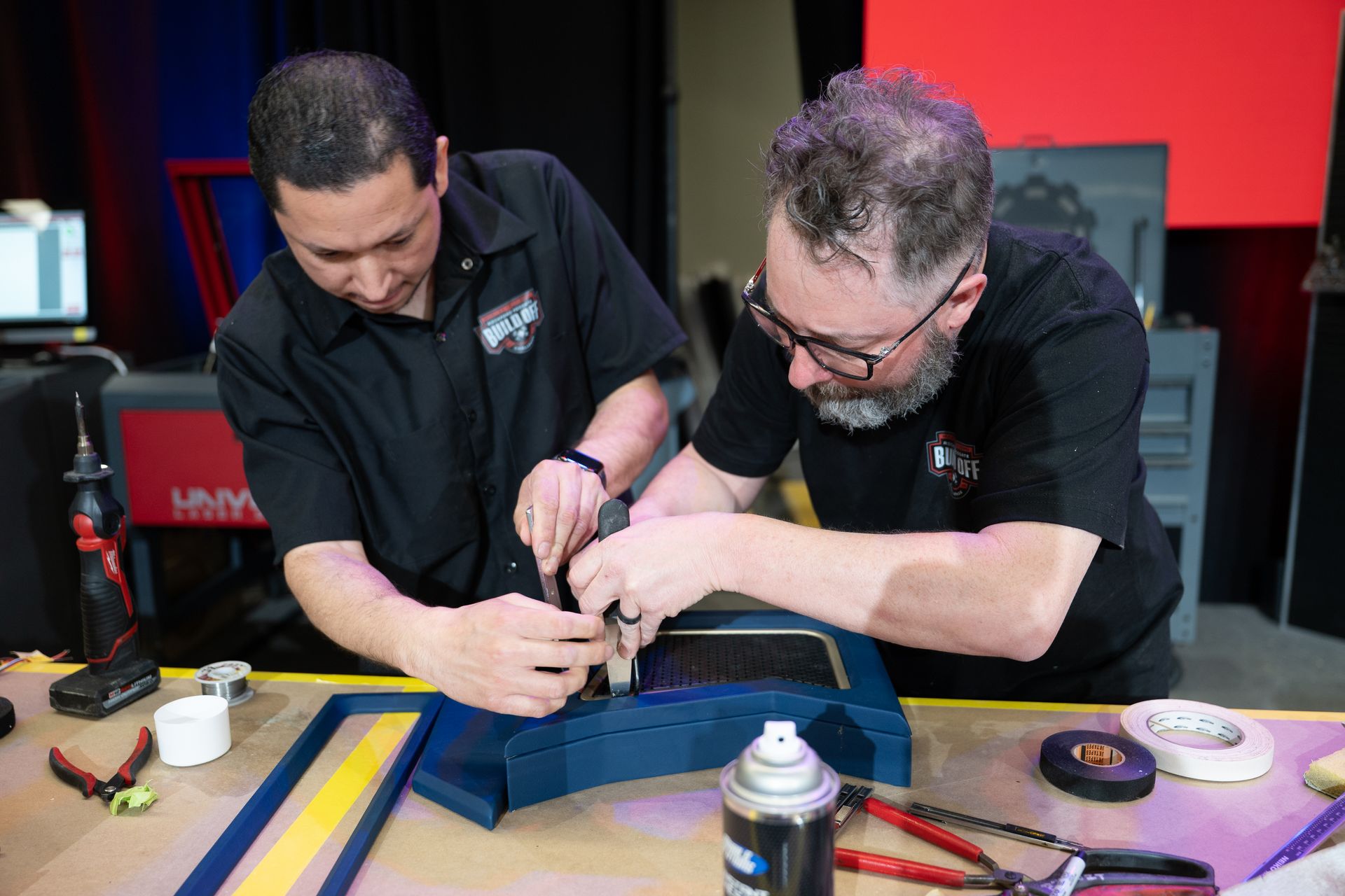Two men working together on a blue car part. They're using tools at a workbench in a workshop.
