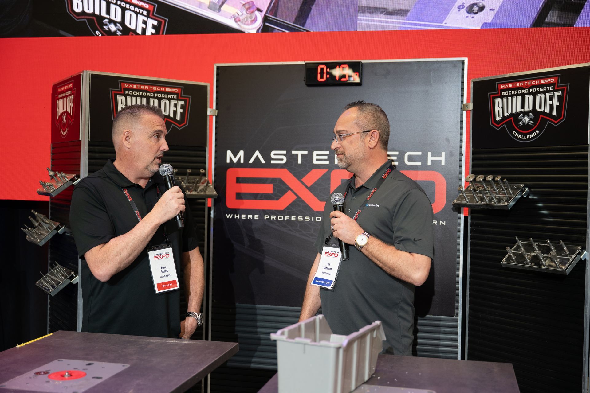 Two men with microphones stand at a trade show booth, likely discussing auto parts.