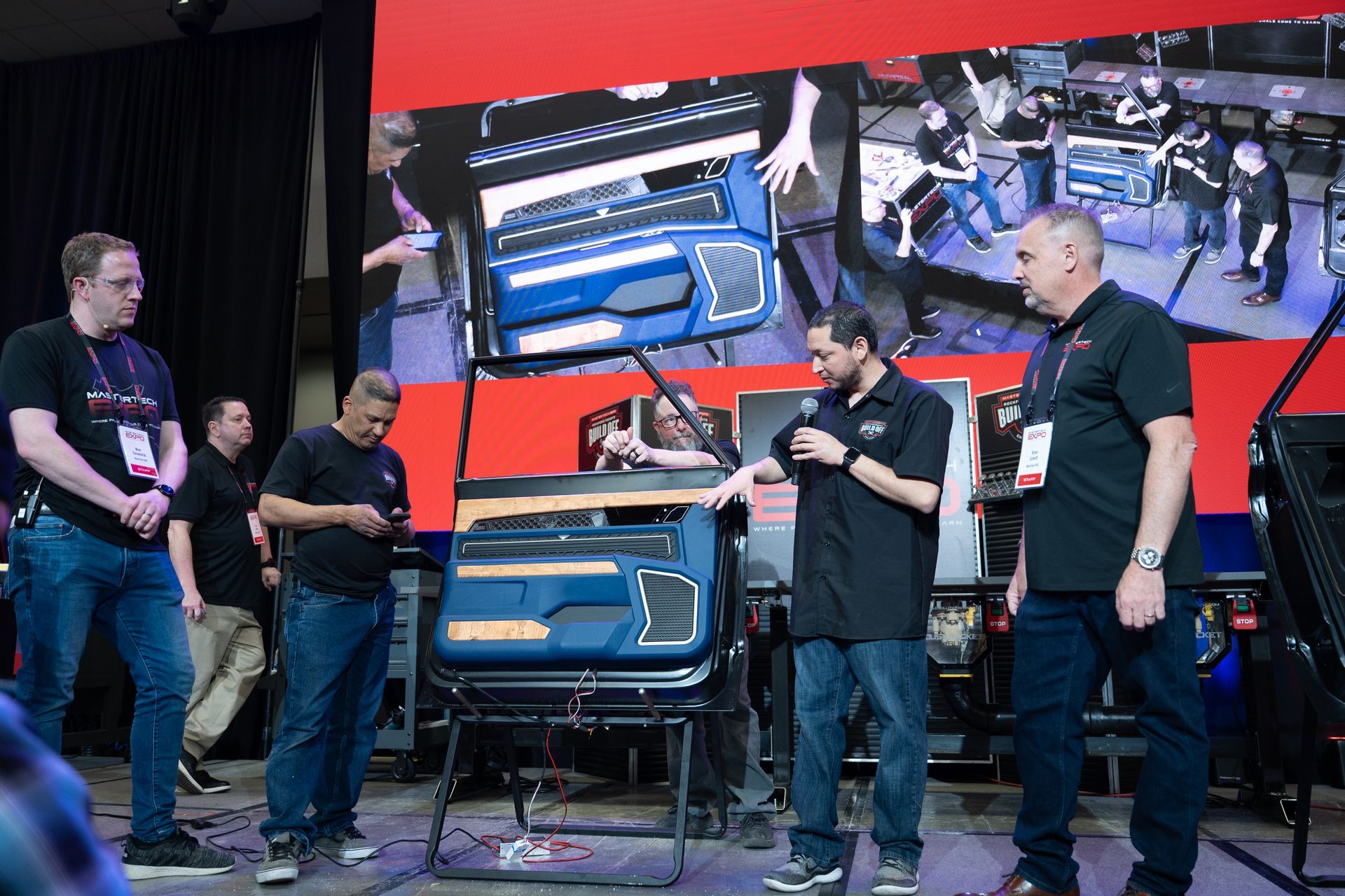 Men demonstrating a blue audio device on a stage with a screen displaying a group.