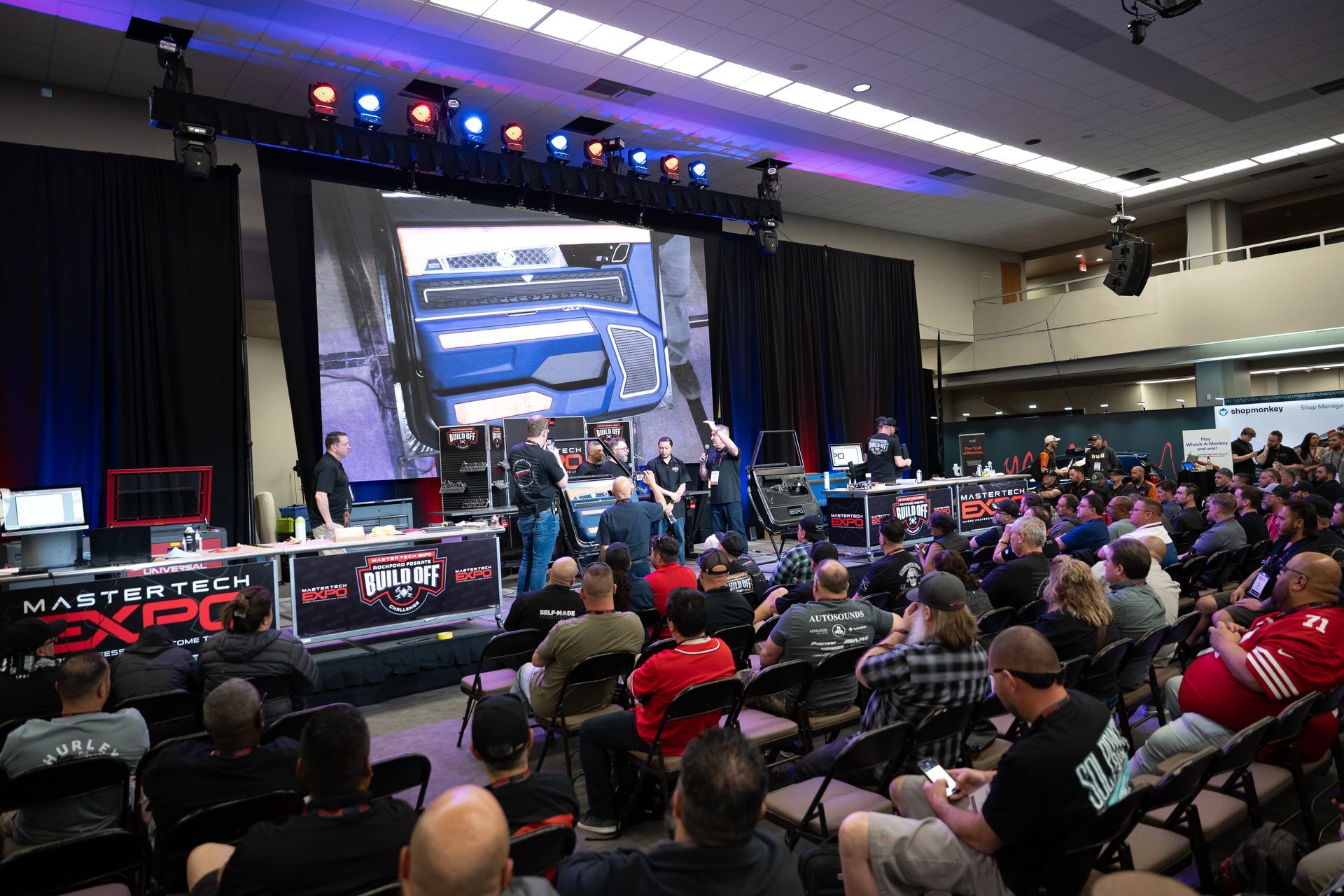 Audience watches a presentation at a technology expo, with a large screen displaying a vehicle.