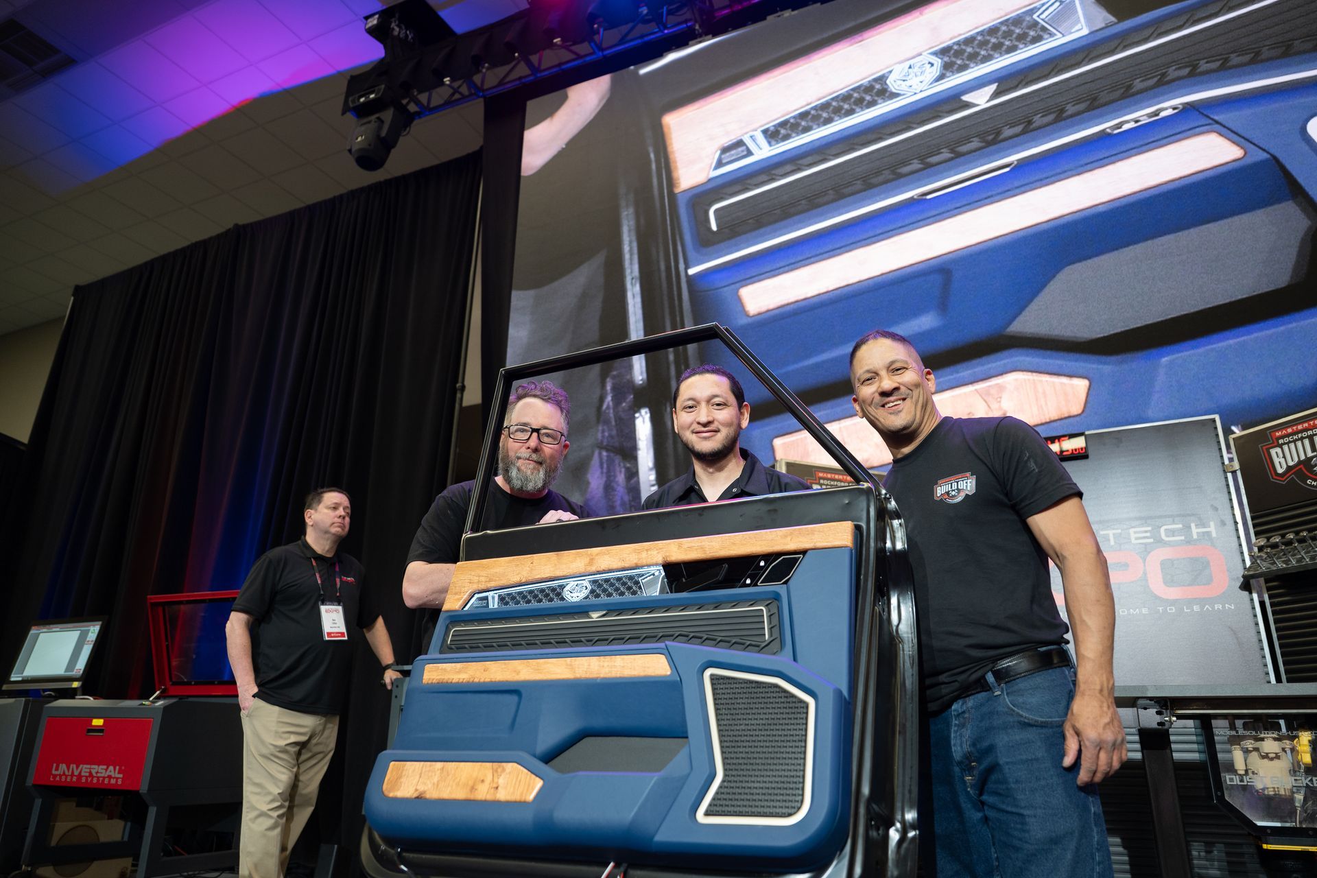 Three men pose with a blue car door at a tech event. One man points, others smile, and a fourth man stands to the side.