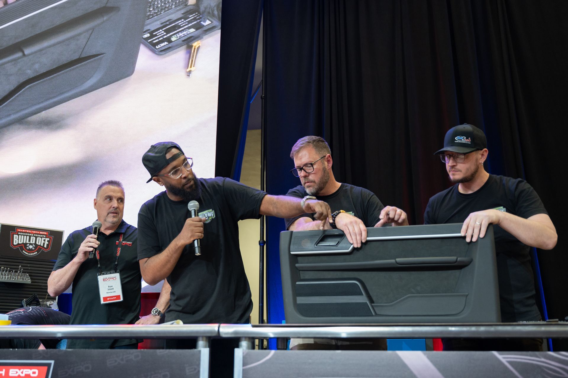Four men at a booth, holding a car door panel, one speaking into a microphone at an expo.