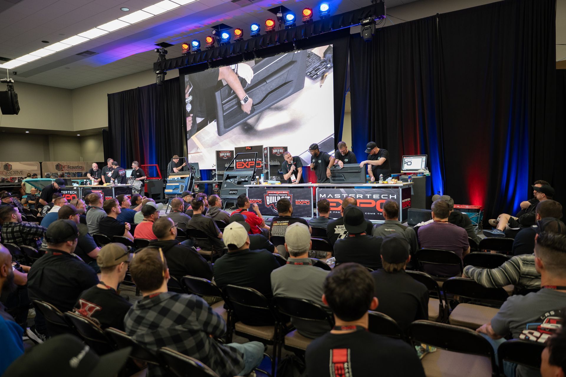 A tech event: audience watching a stage with a large screen, people assembling equipment, and stage lighting.