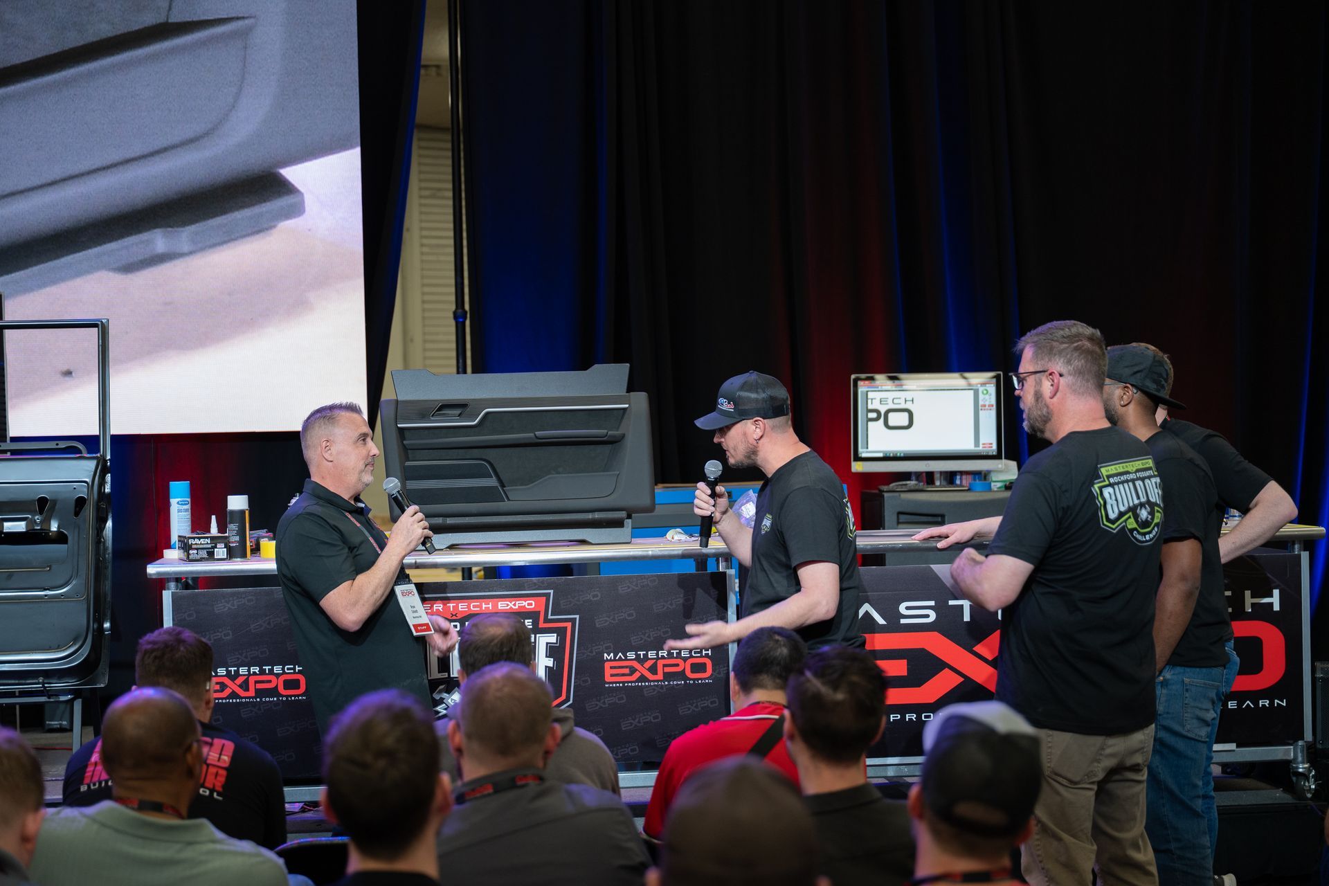 People on stage at a convention talking near car interior parts. A crowd watches.