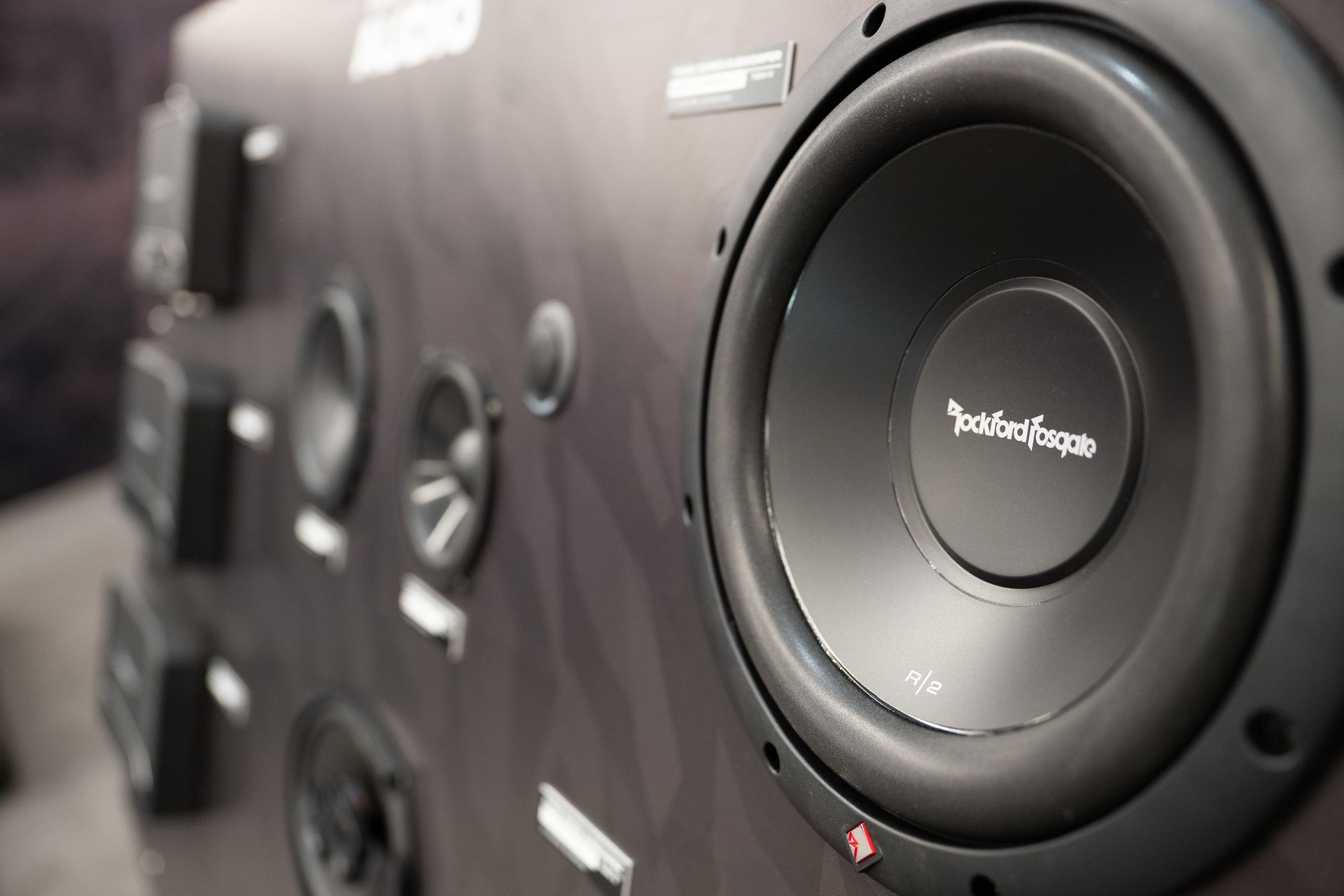 Close-up of a black Rockford Fosgate subwoofer on a display panel with other car audio speakers.