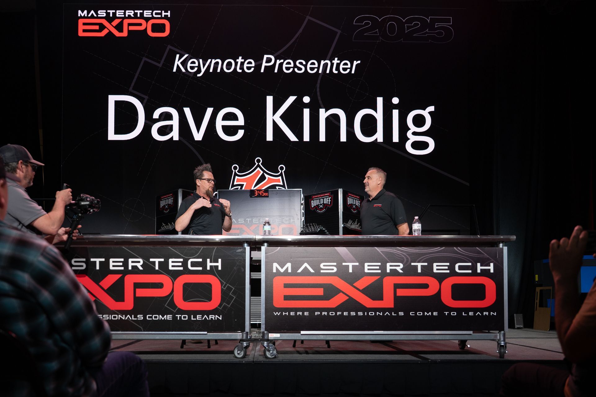 Dave Kindig speaking at the MasterTech Expo. Black backdrop, two men seated at a table. Red and white logo.