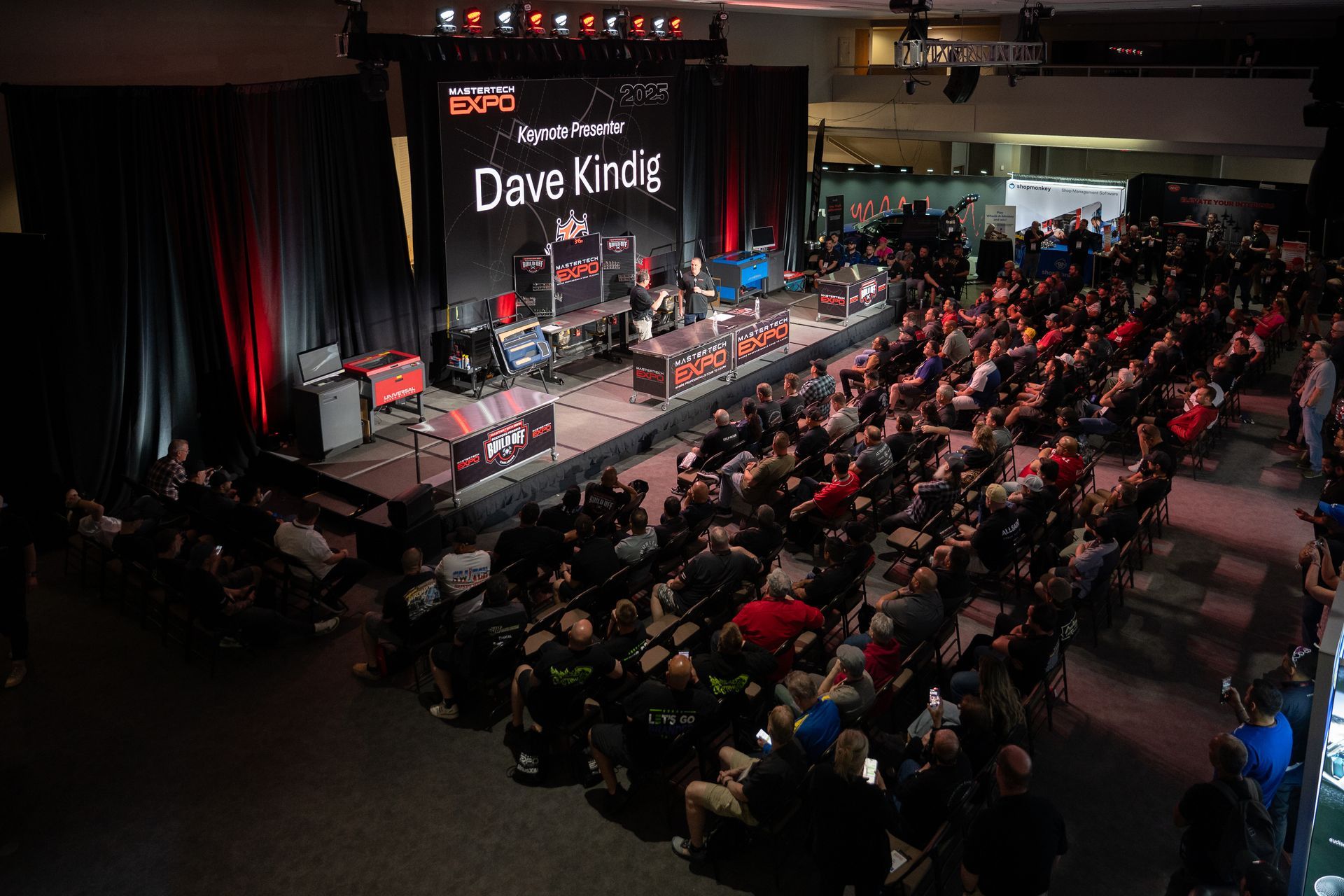 A large audience watches a presentation on a stage; a screen displays 
