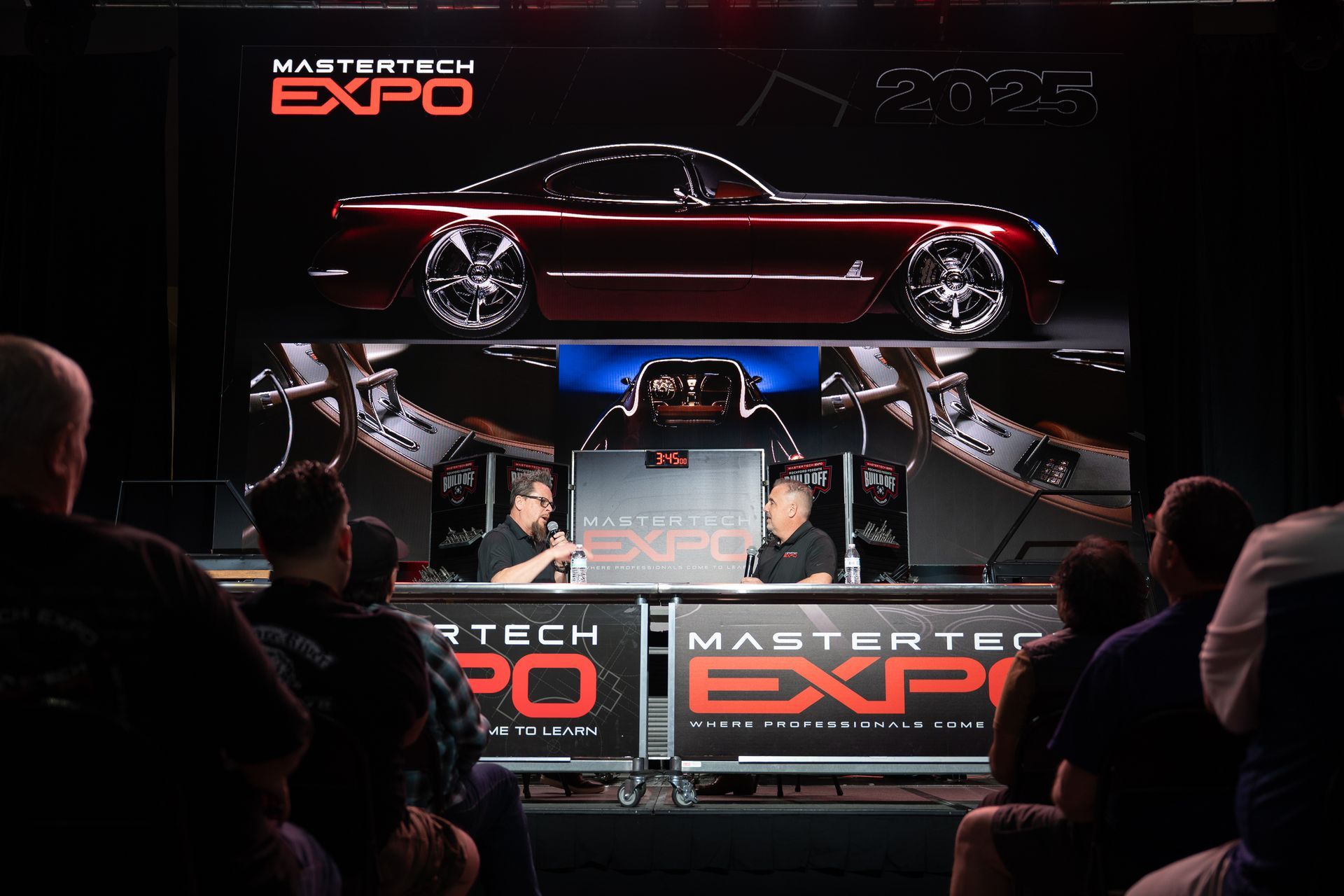 MasterTech Expo: Two men on stage, a lifted maroon car on display, audience in attendance.