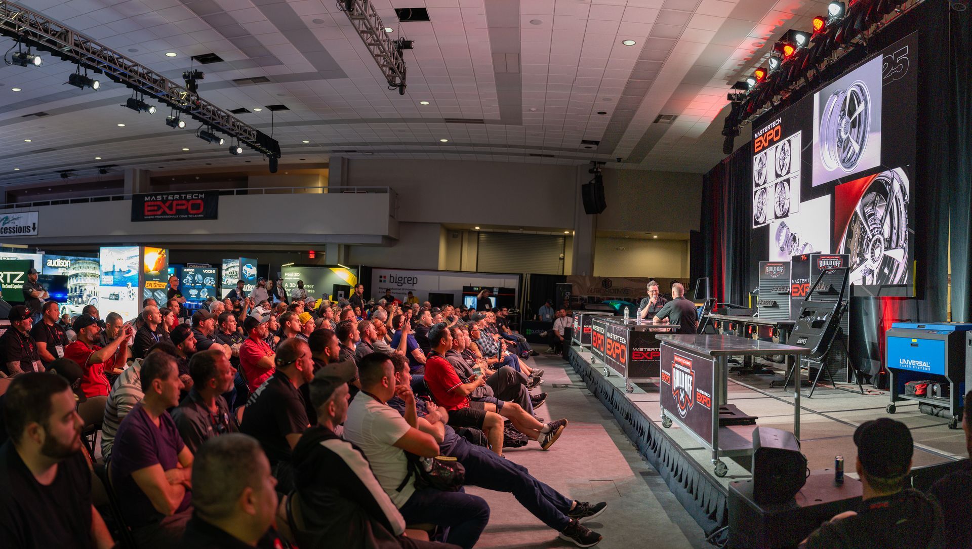 Audience watches panel discussion on stage at event, with large screen, red lighting, and exhibits in background.