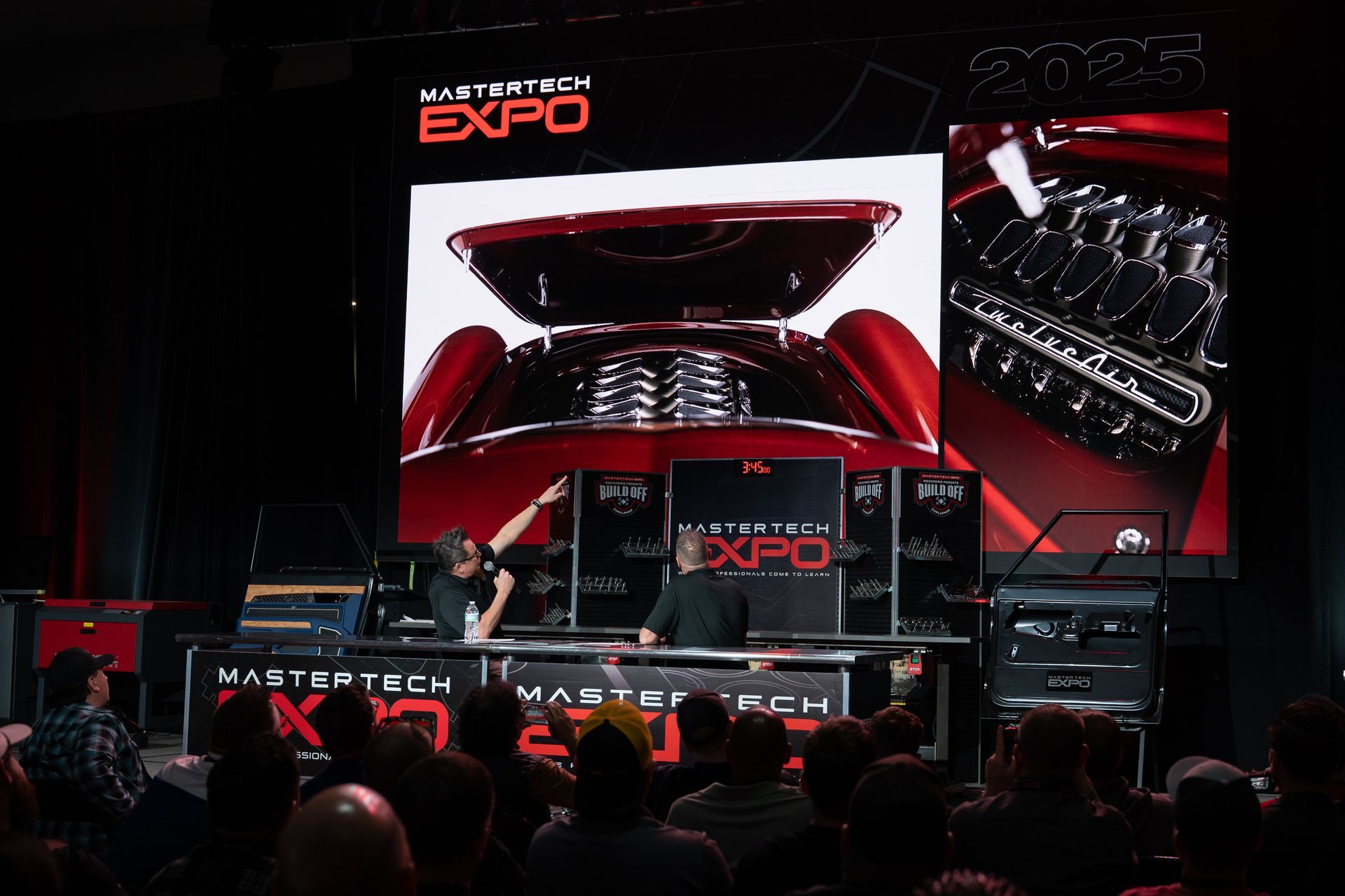 A presenter at MasterTech Expo points to a car engine on screen; a crowd watches.