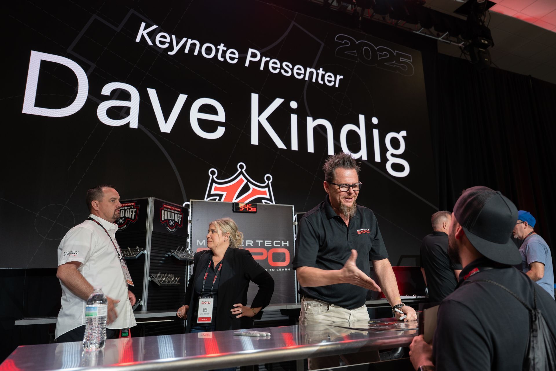 Dave Kindig, Keynote Presenter, speaks at a conference behind a table with staff in a large venue.