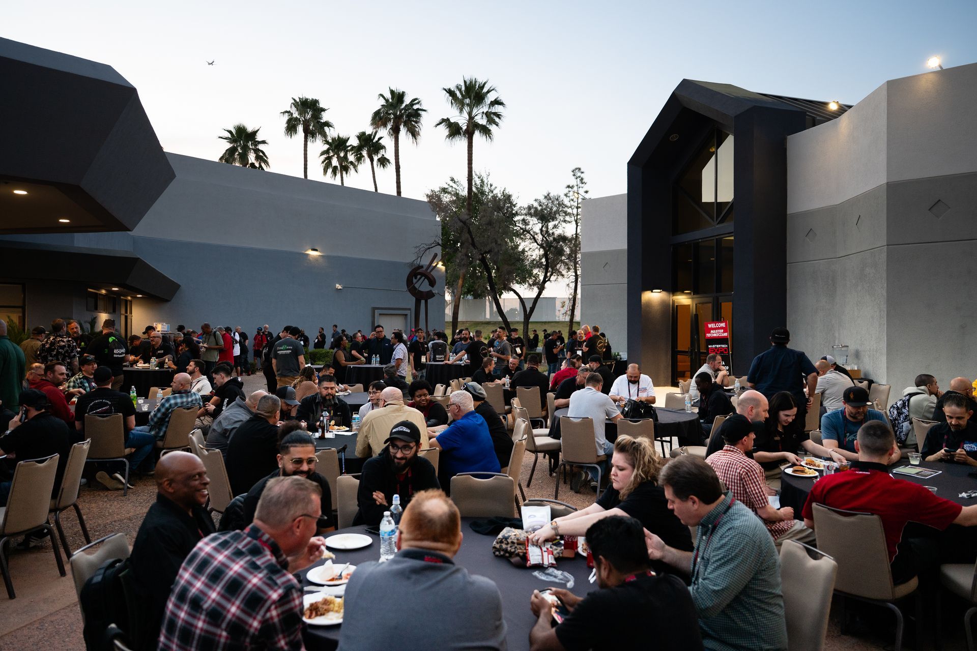 Large outdoor gathering at tables, eating and socializing in front of a building with palm trees.