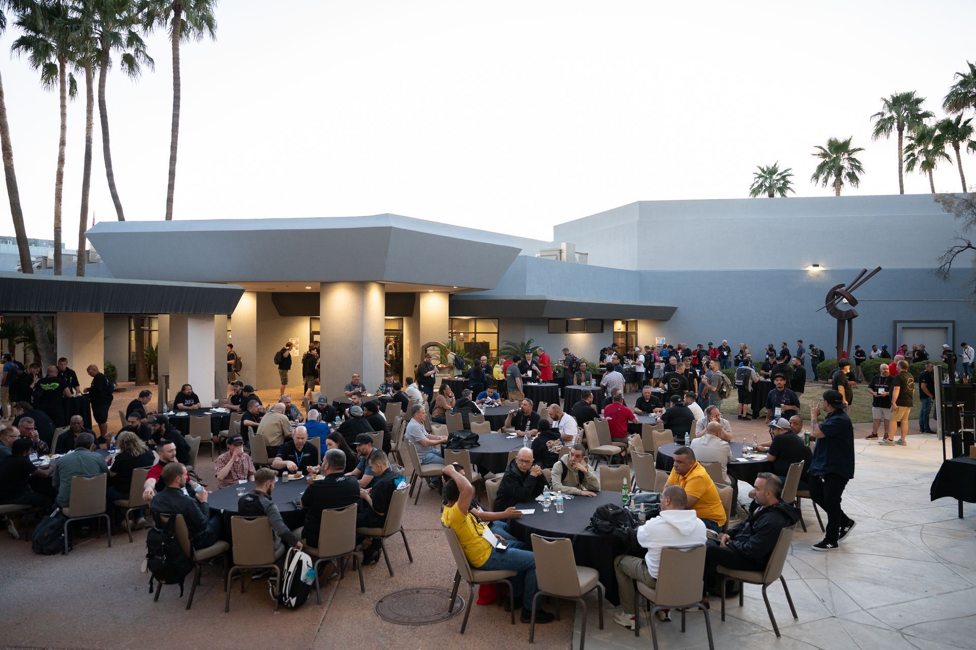 Outdoor event with people seated at round tables. A building and palm trees are in the background.