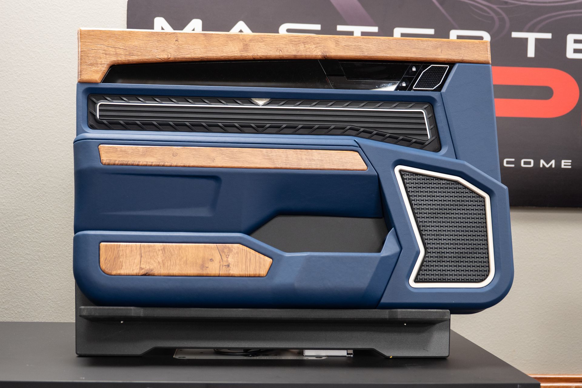 Blue and wood-grain car door panel on a black display stand. Interior car detail with speaker grille and controls.