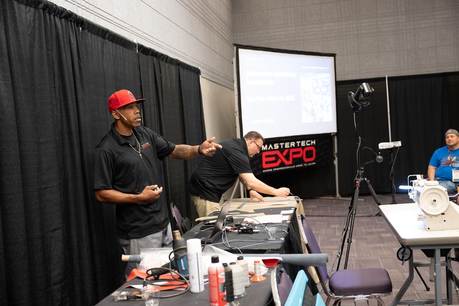 Man in red hat gesturing during a presentation at a conference. Others work at a table with equipment.