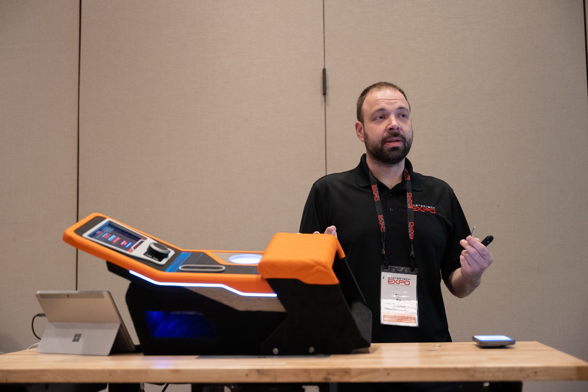 Man presenting at a table with an orange and black machine; neutral background.